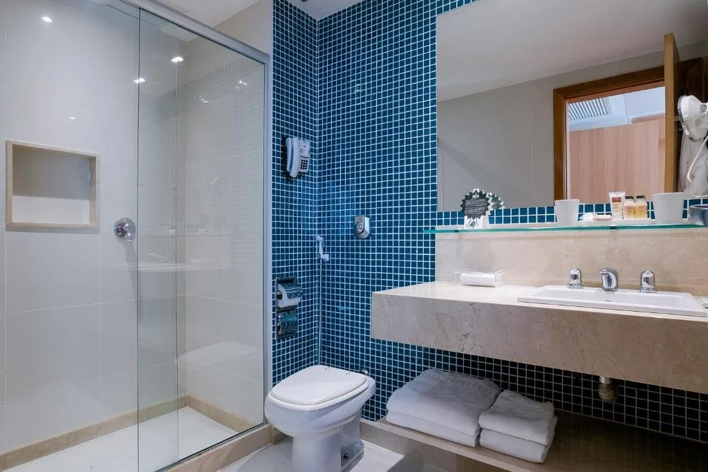 Bathroom with a glass shower enclosure on the left, a white toilet in the middle, and a beige marble vanity with a sink and a large mirror on the right. Blue mosaic tiles cover the wall behind the toilet and vanity. Bathroom toiletries and towels are visible.