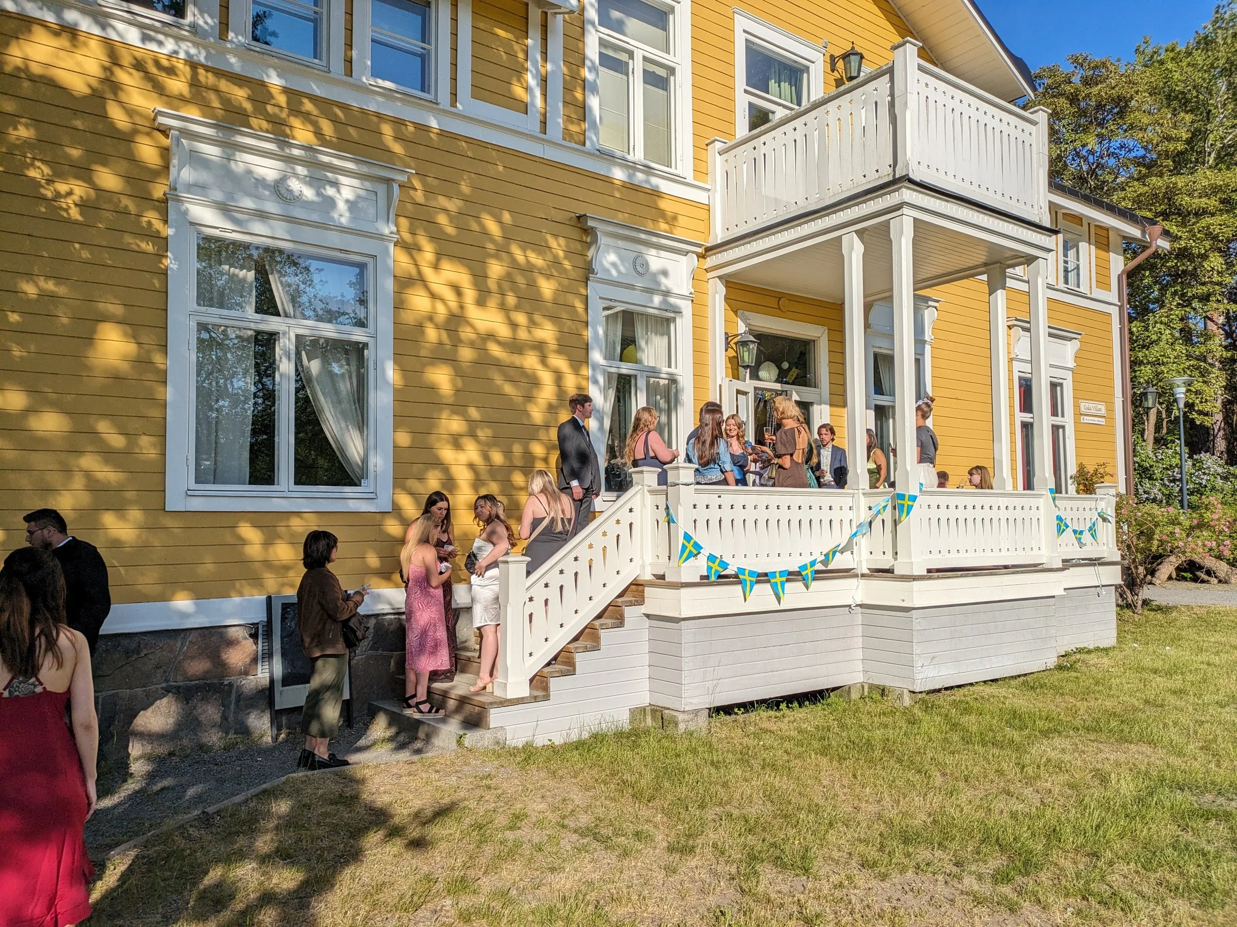 People gathered on the porch and steps of a yellow house with white window frames and a small balcony, possibly celebrating a Swedish event, with decorative blue and yellow banners.