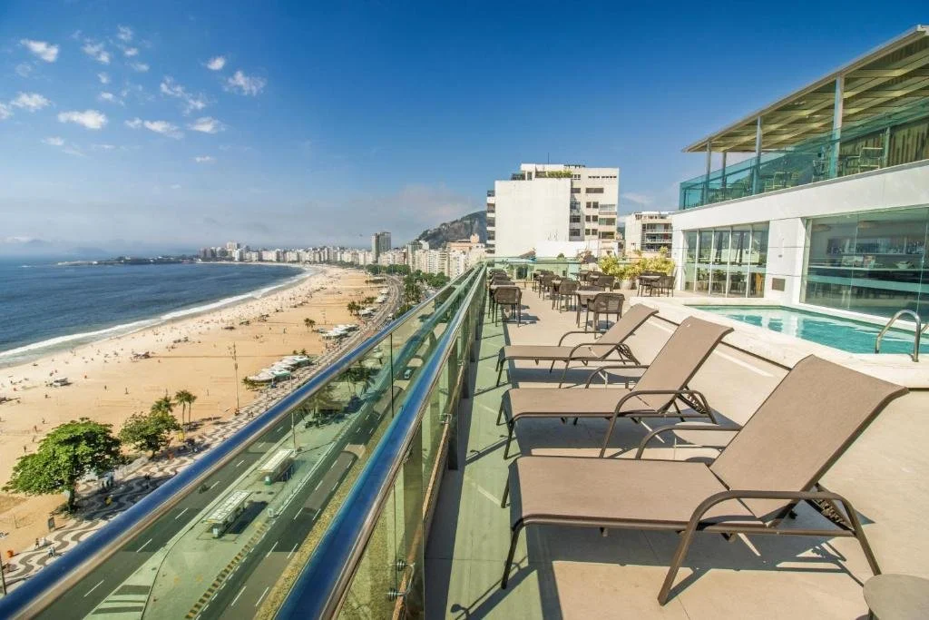 A balcony with lounge chairs overlooking a sandy beach and ocean with a city skyline in the distance.