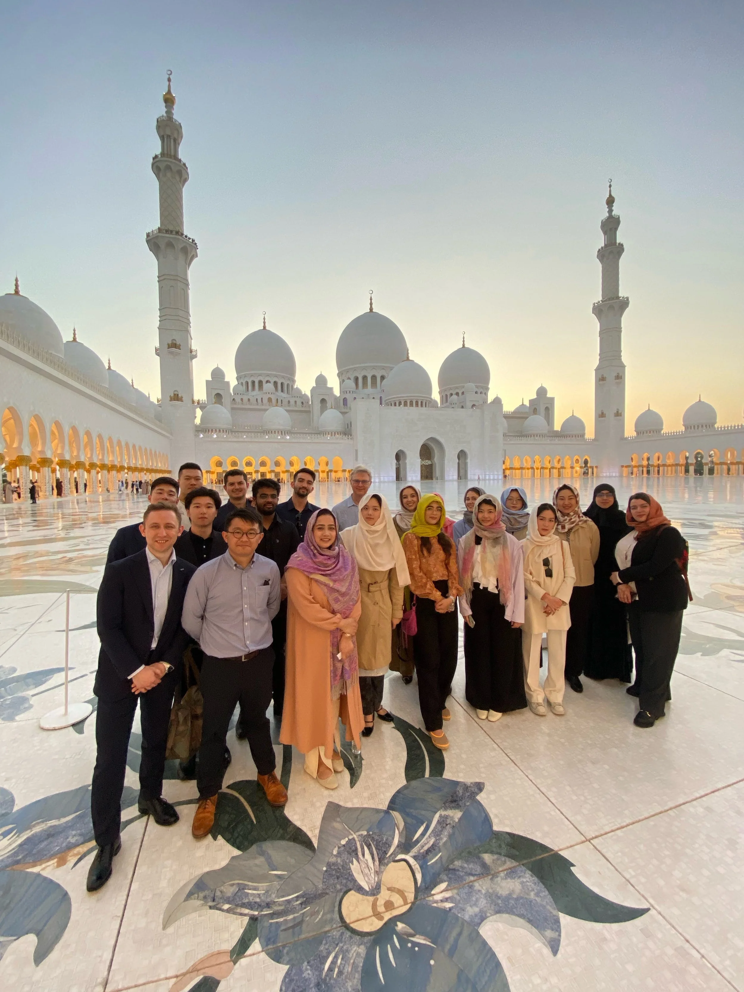 Group of people standing in front of the mosque in the courtyard during sunset, with domes and minarets in the background.