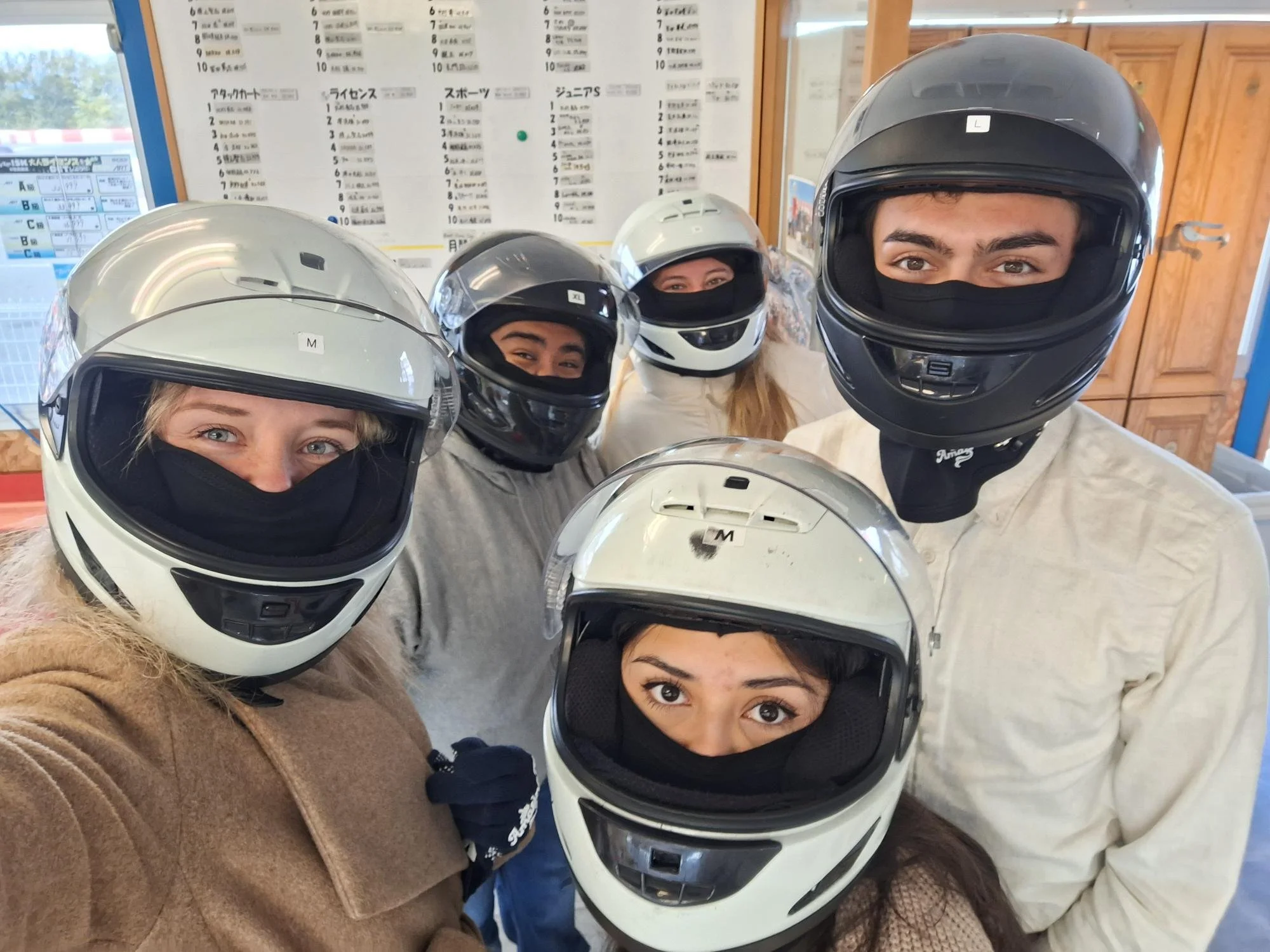 Group of five people wearing motorcycle helmets indoors, some with their faces partially covered with masks, smiling and looking at the camera.