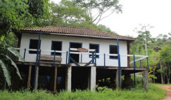 A small house on stilts with a brown tiled roof, white walls, and a wooden porch surrounded by greenery.