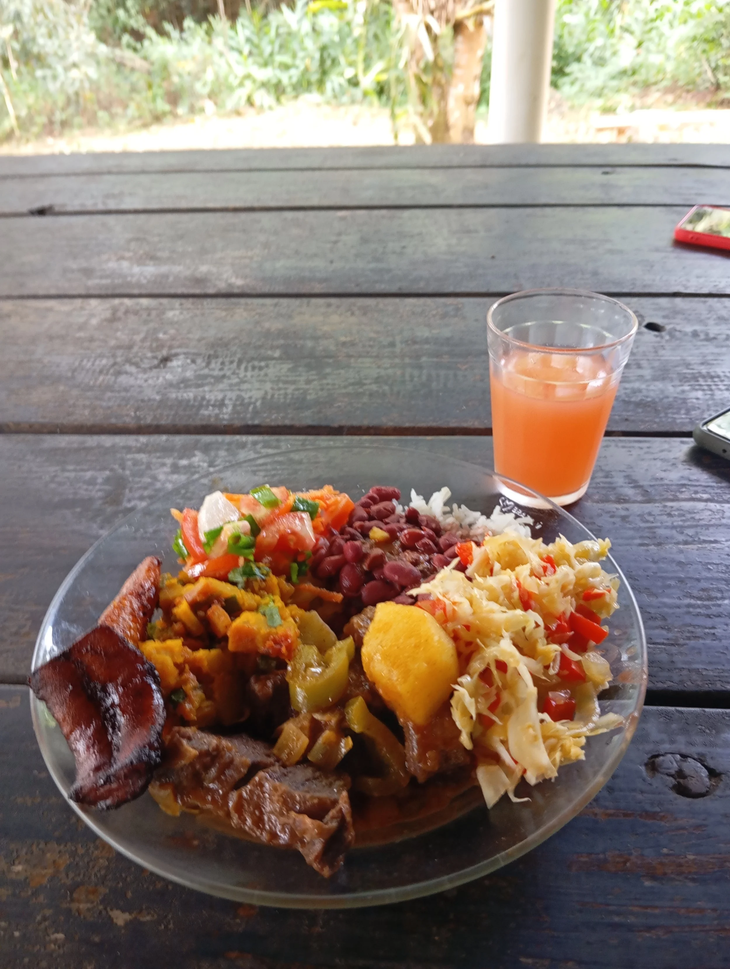 Plate of traditional Latin American food with various bean and vegetable dishes, a glass of pink juice, and a partially visible mobile phone on a wooden table outdoors.