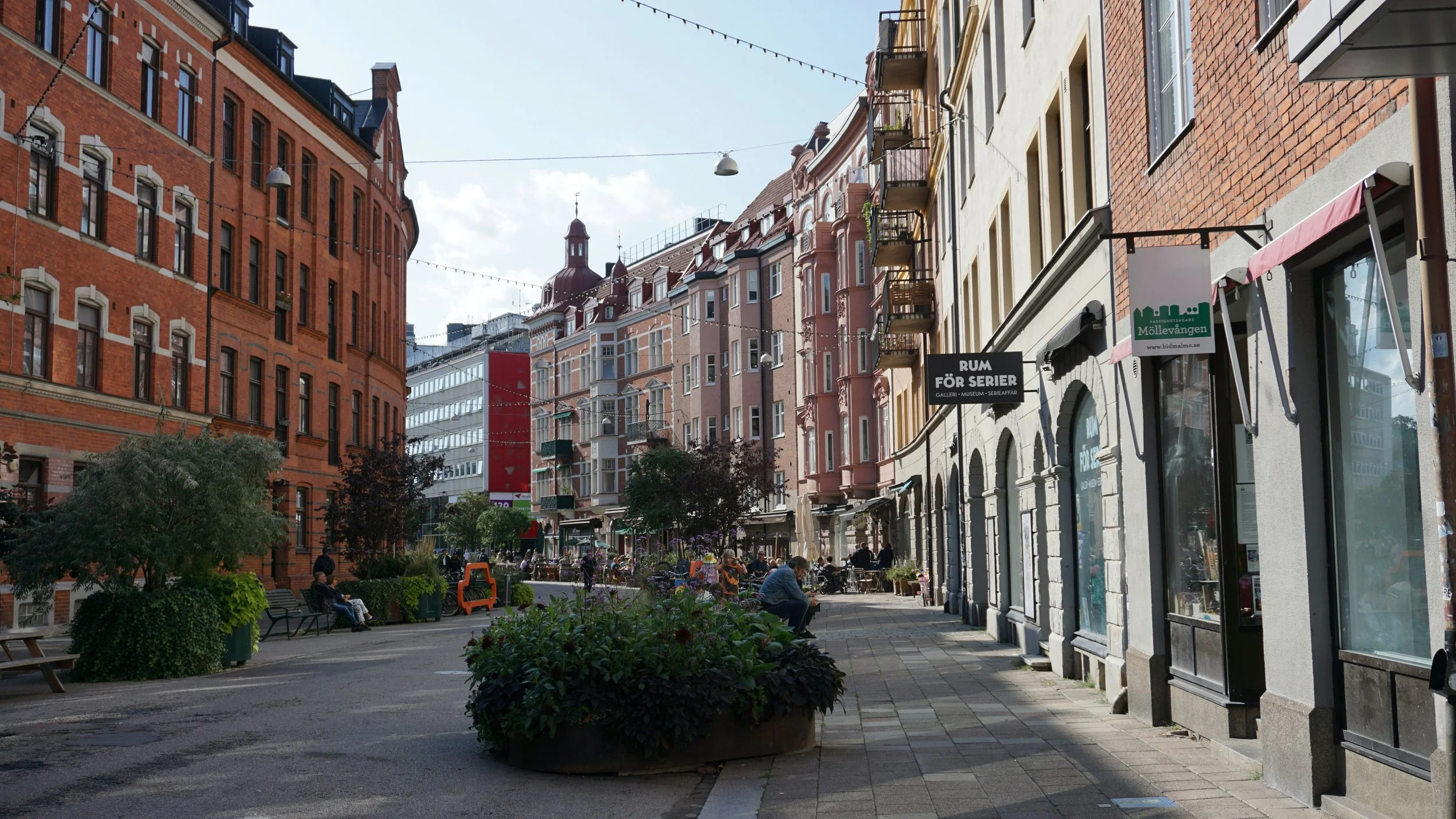 A city street with colorful historic buildings, sidewalk cafes, benches, trees, and pedestrians.