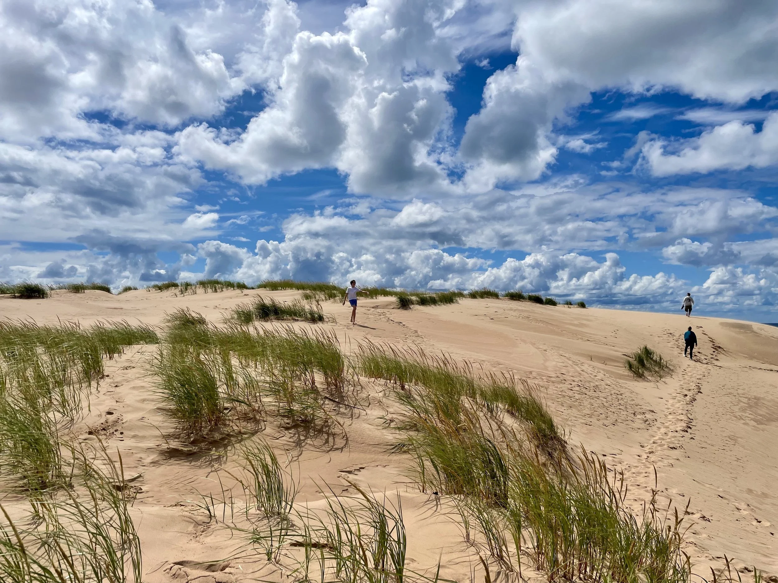 People walking on sandy dunes with scattered grass, under a partly cloudy sky.