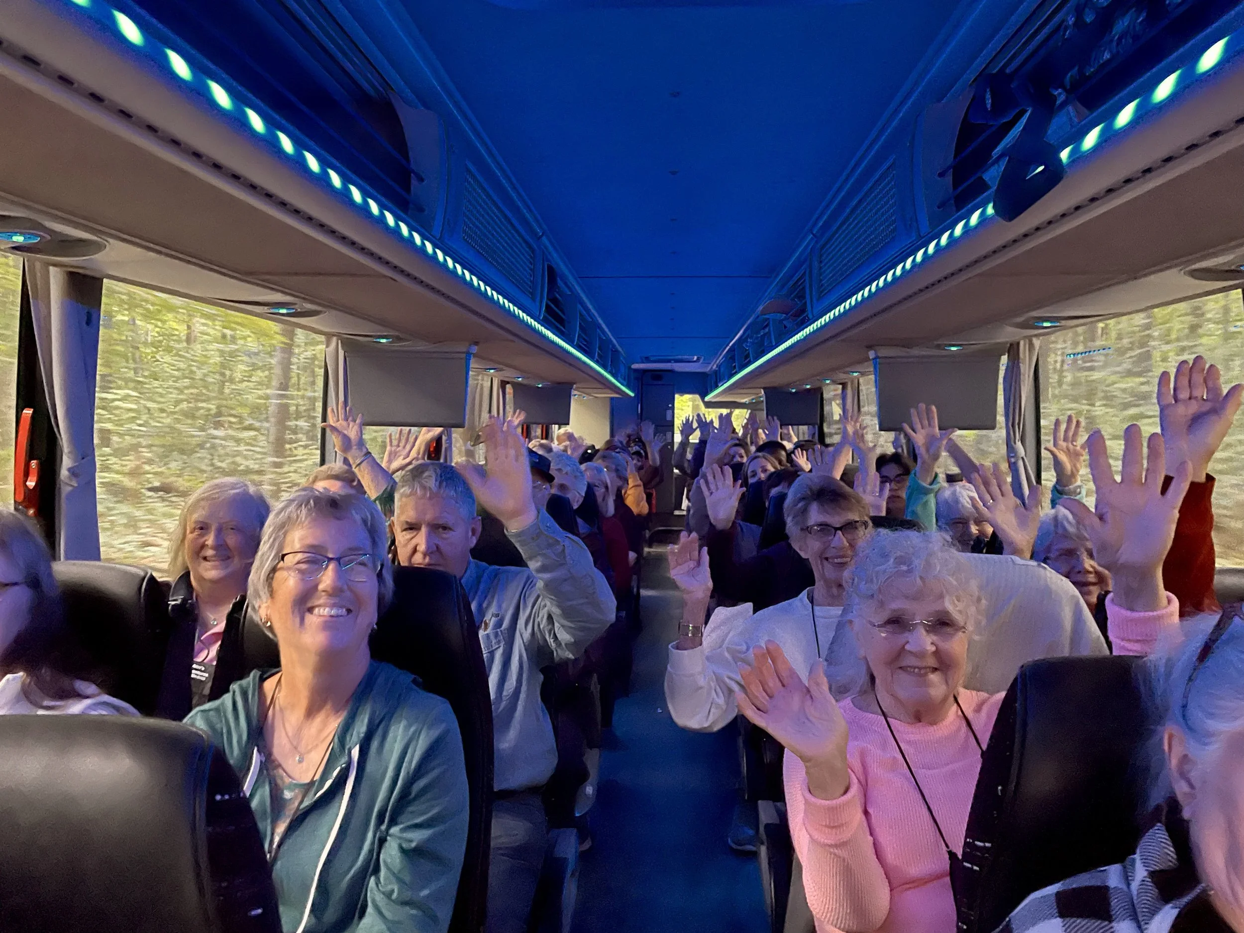 Smiling seniors on a bus, waving at the camera with a forest passing by outside the windows.