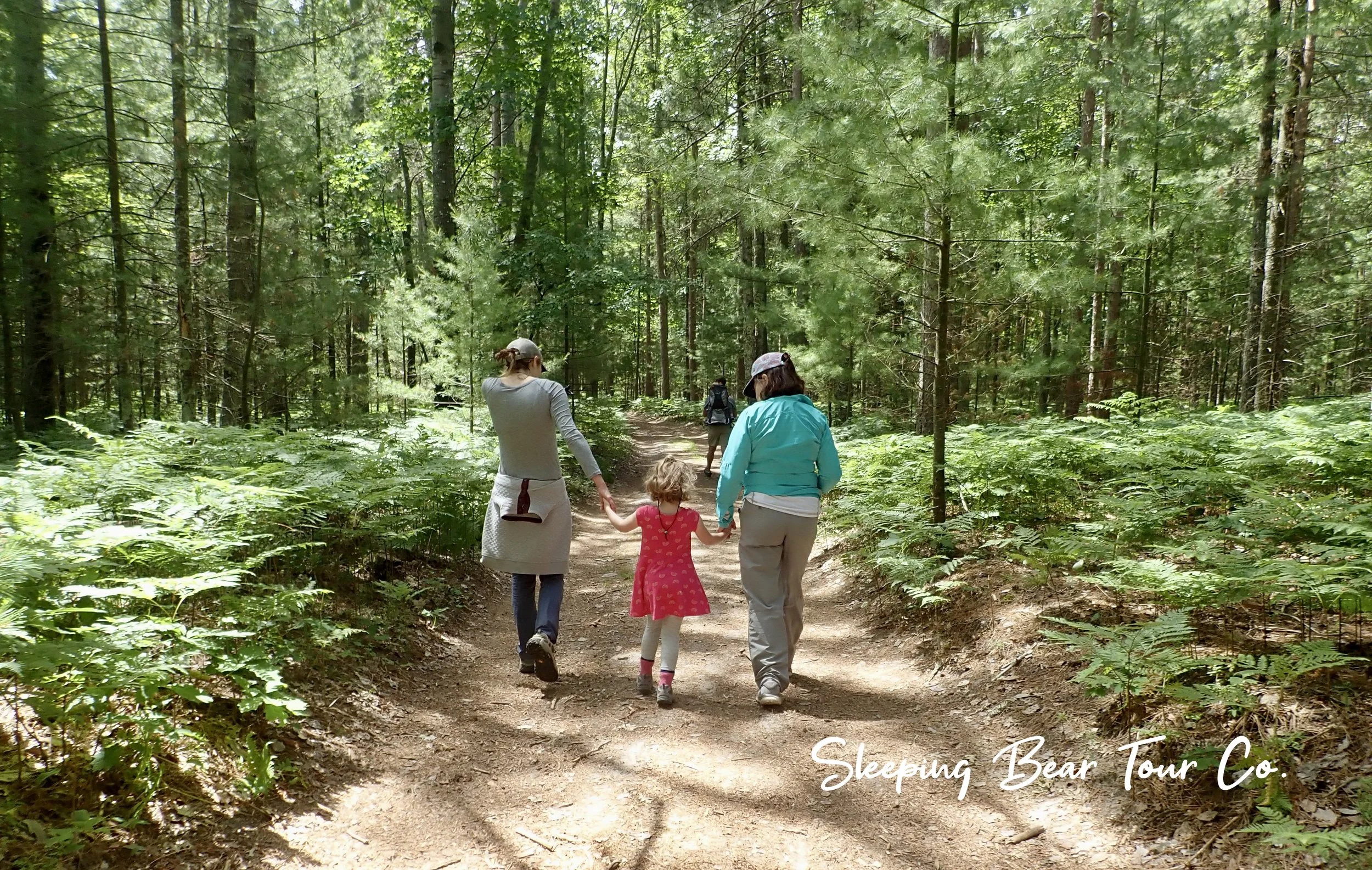 Three people, two adults and a child, walking on a wooded trail surrounded by greenery during daytime. The woman on the left and the woman on the right are holding hands with the young girl in the middle, who is wearing a red dress.