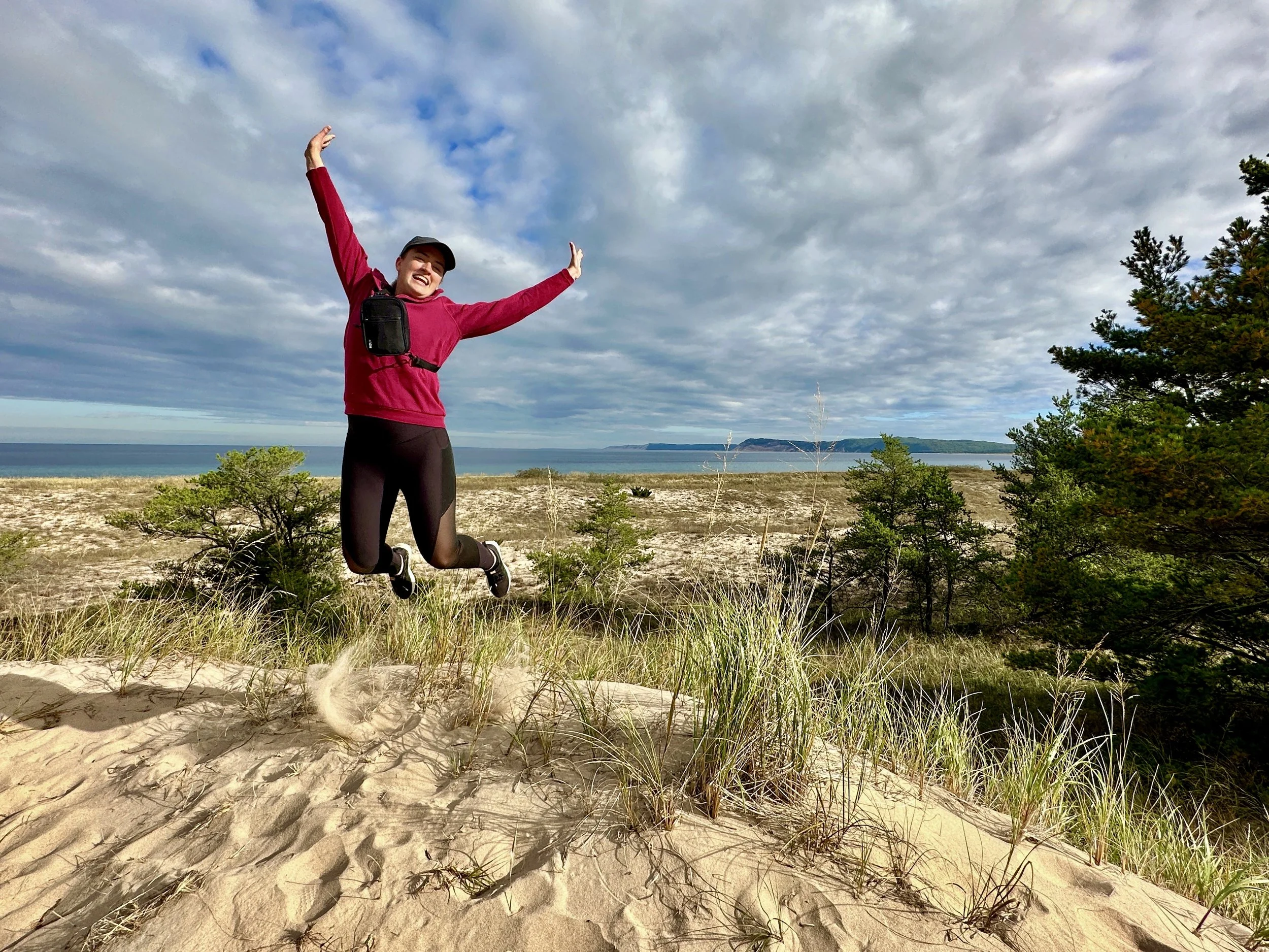 Person jumping on sand dune with trees, Lake Michigan, and cloudy sky in the background