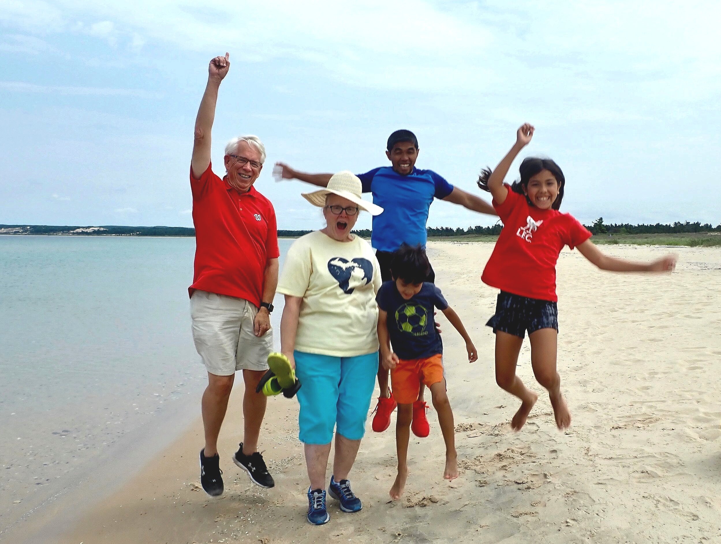 Group of six people, three adults and three children, jumping and smiling on a beach with sand and water, under a partly cloudy sky.