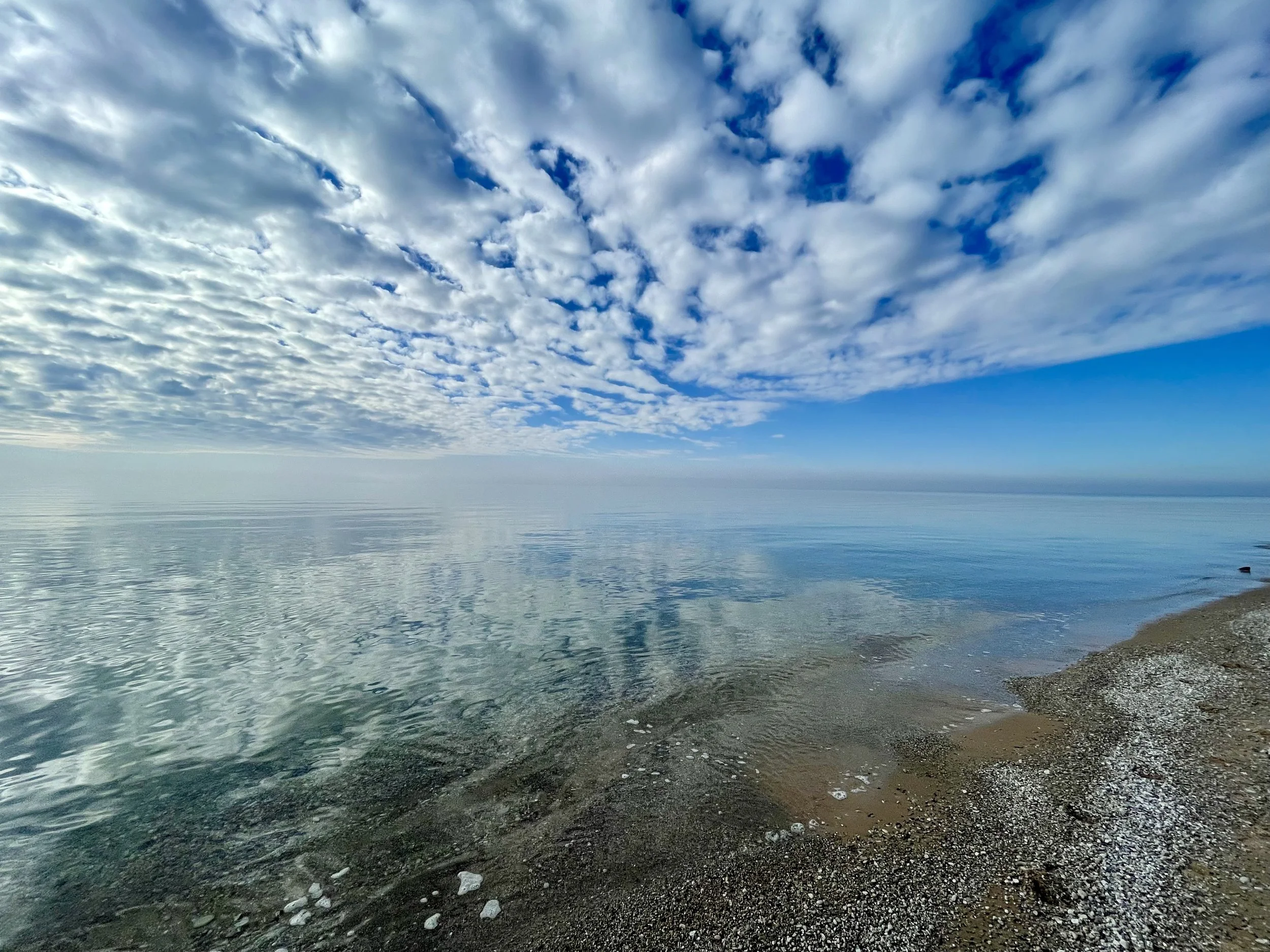 Serene seashore with calm water reflecting a sky filled with scattered clouds on a clear day.