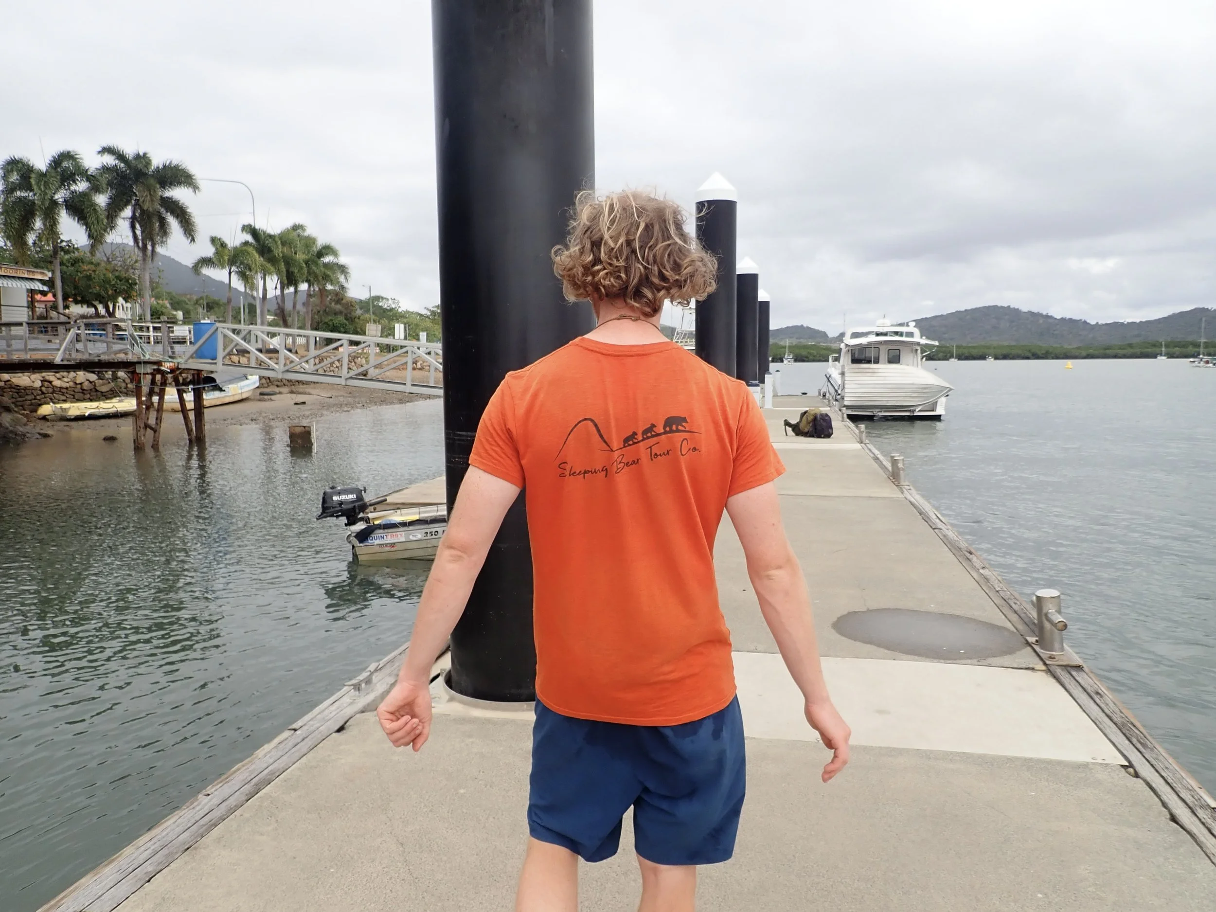 A person with curly hair wearing an orange t-shirt and blue shorts walks on a pier toward boats and water on a cloudy day.