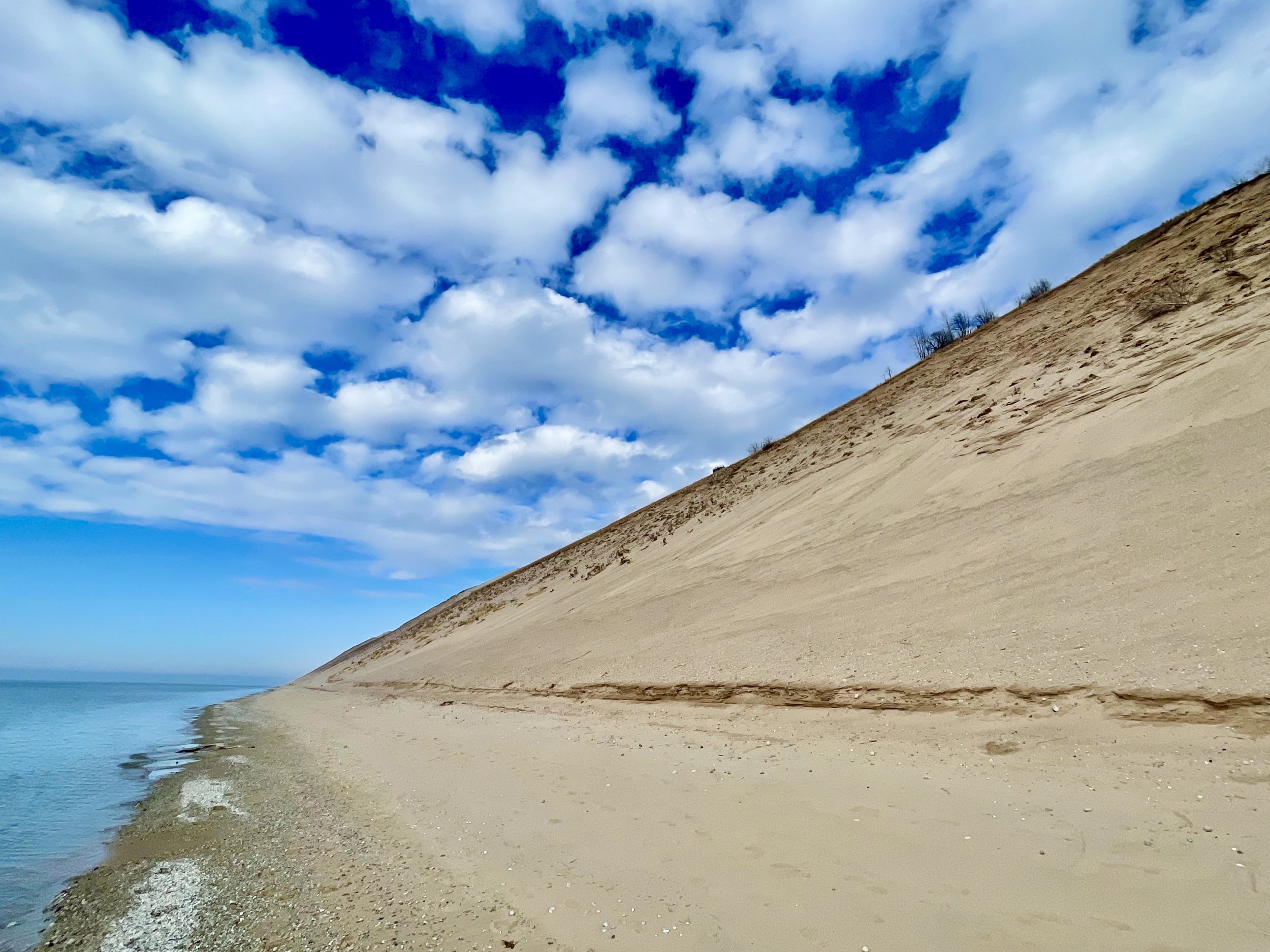 A sandy beach with a large sand dune on the right, calm water on the left, and a blue sky with scattered clouds overhead.