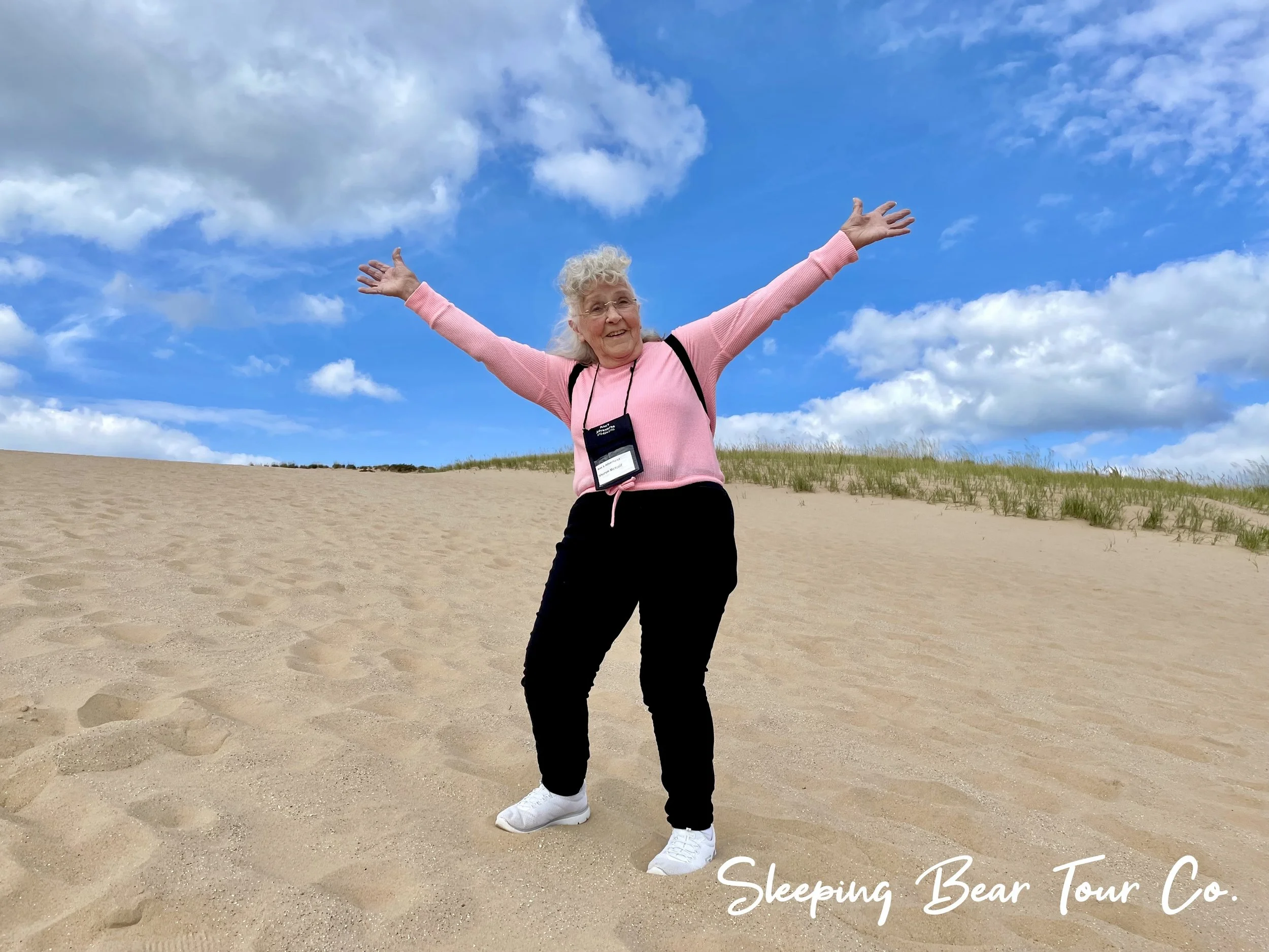 A joyful elderly woman with curly gray hair in a pink sweater and black pants standing on a sandy hill with arms outstretched under a partly cloudy blue sky. She wears white sneakers and has a black camera around her neck. The text "Sleeping Bear Tour Co." is in the bottom right corner.