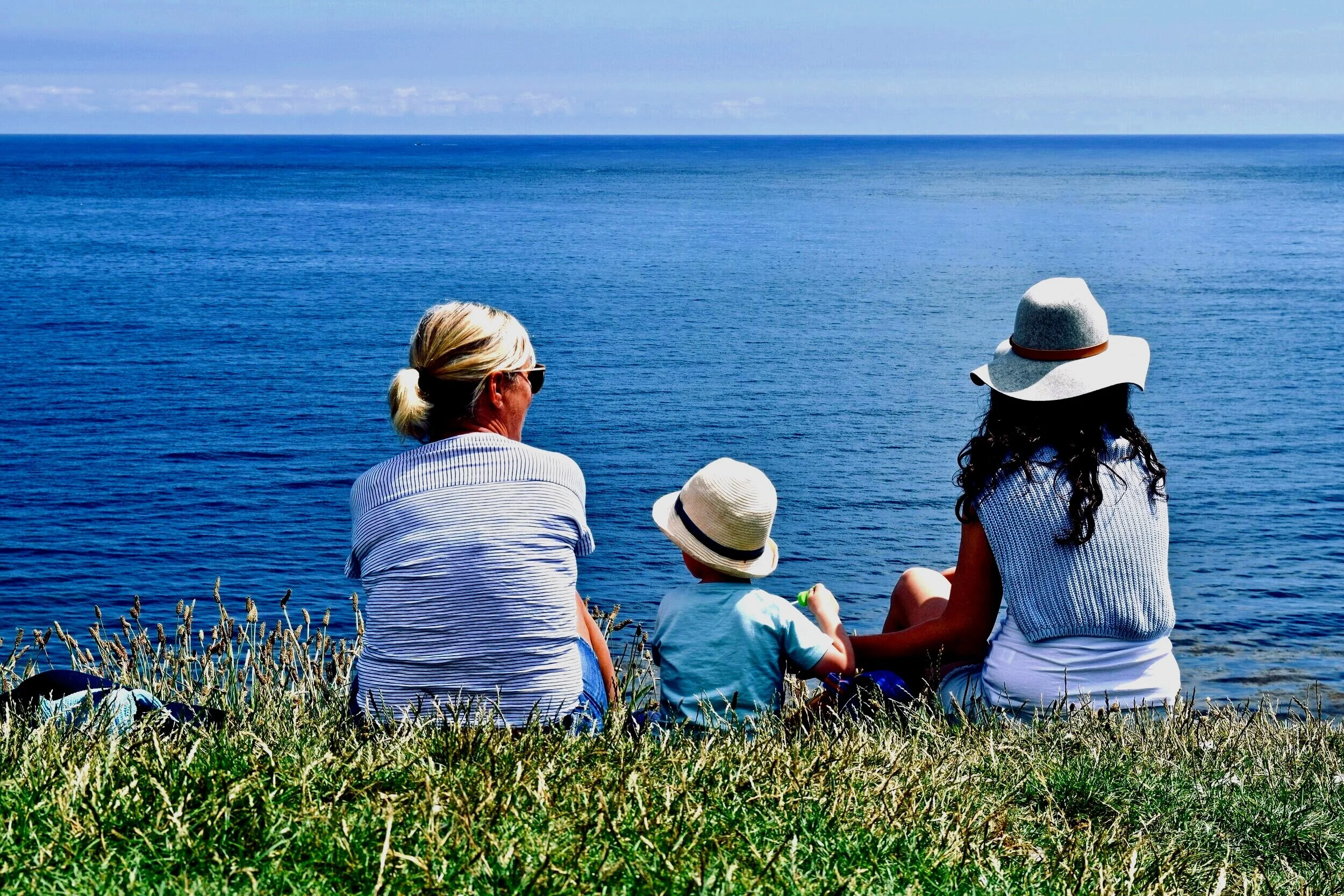 Three people, two women and a child, sitting on grass by the water and facing Lake Michigan on a sunny day, wearing hats and sunglasses.