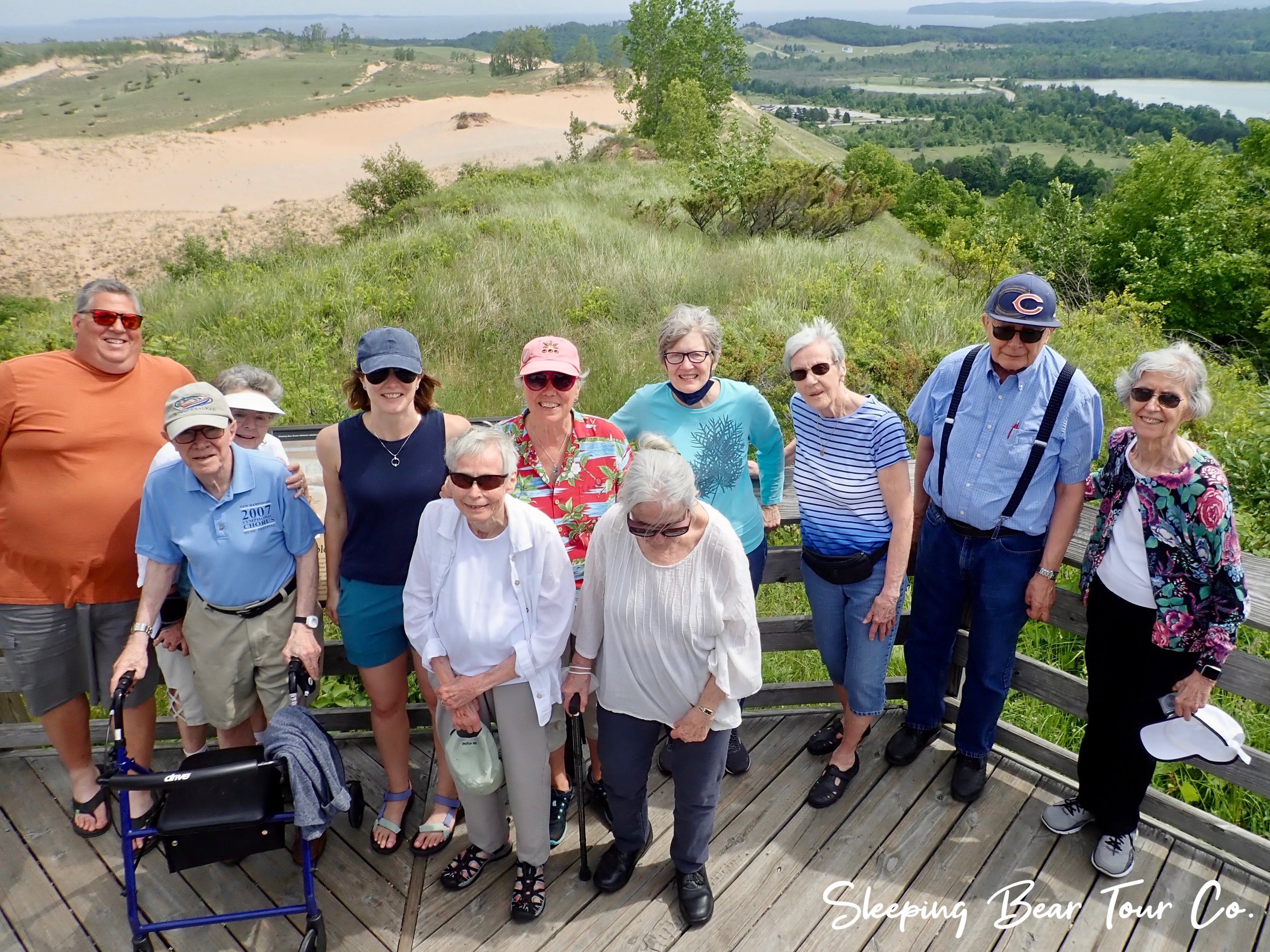 Group of people posing on a wooden deck with a scenic view of dunes and lush greenery in the background, wearing casual summer clothing and sunglasses.