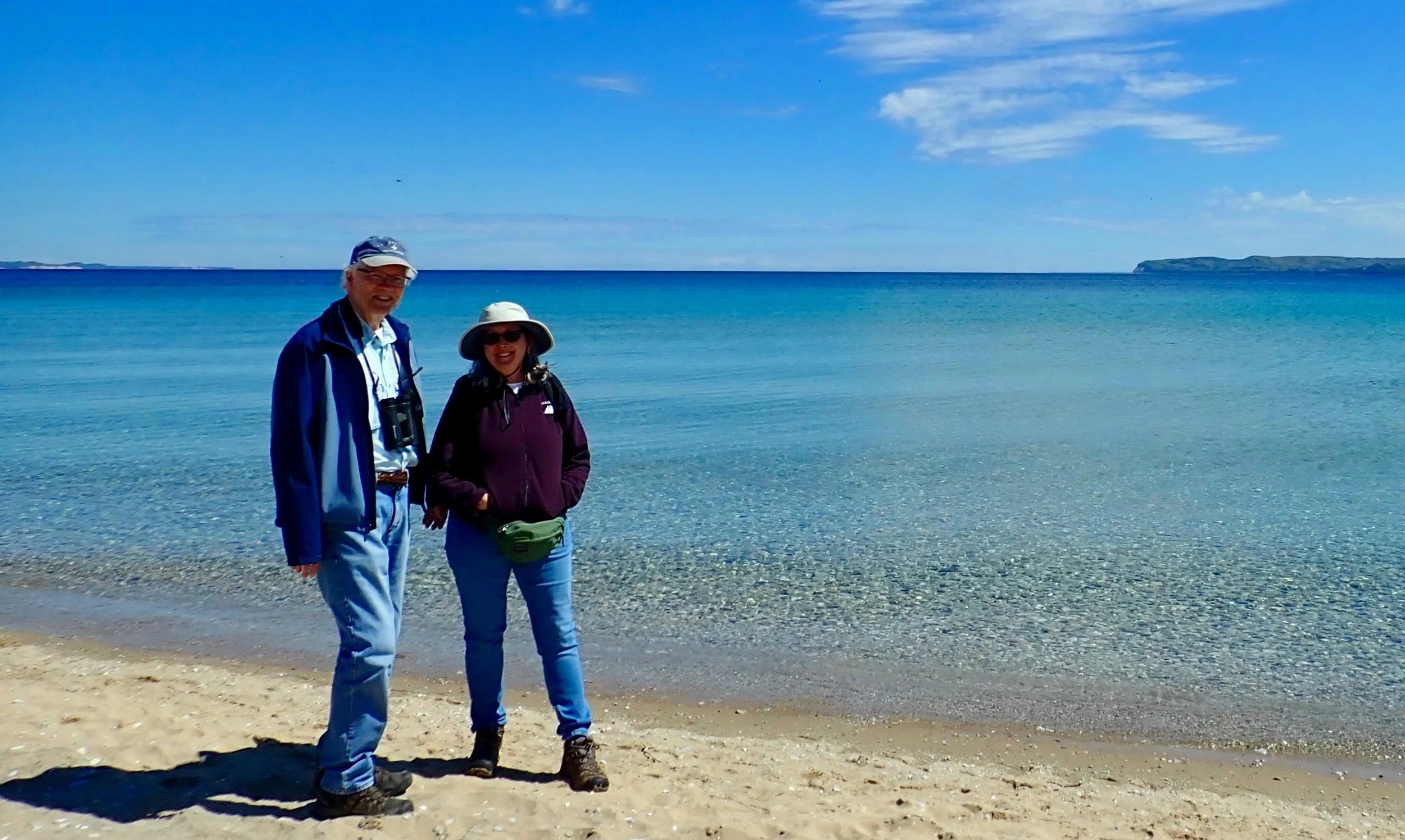 Two people standing on a sandy beach with a clear blue sky and ocean background.