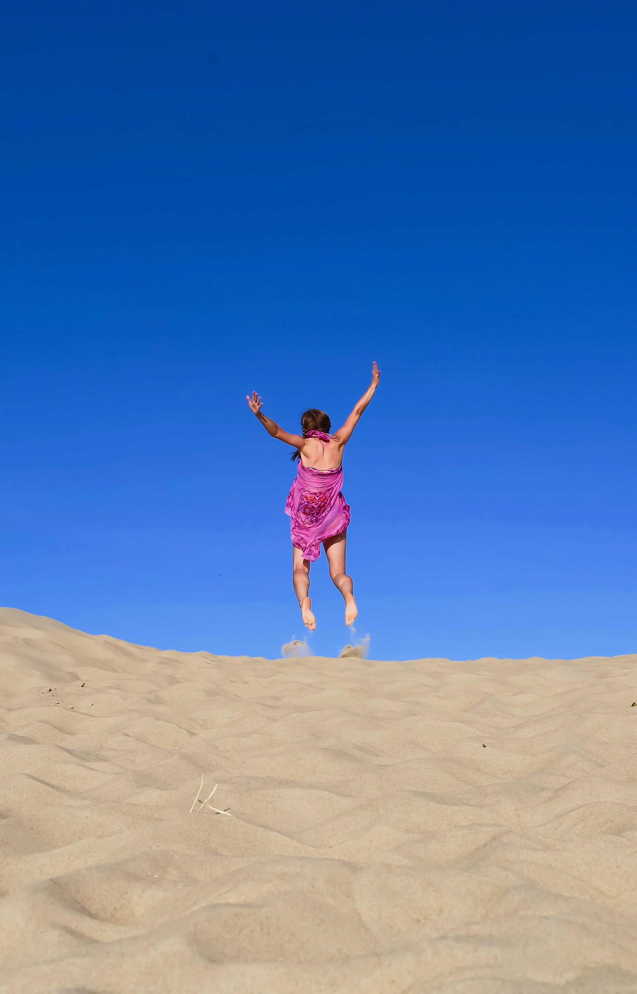 A girl in a pink dress jumping in the air on a sandy beach with a clear blue sky background.