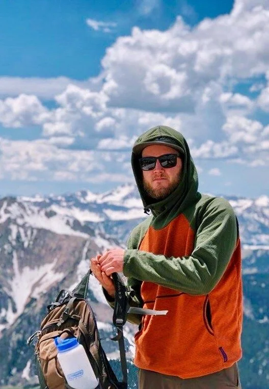 Person in hooded jacket and sunglasses standing on snowy mountain with backpack and water bottle.