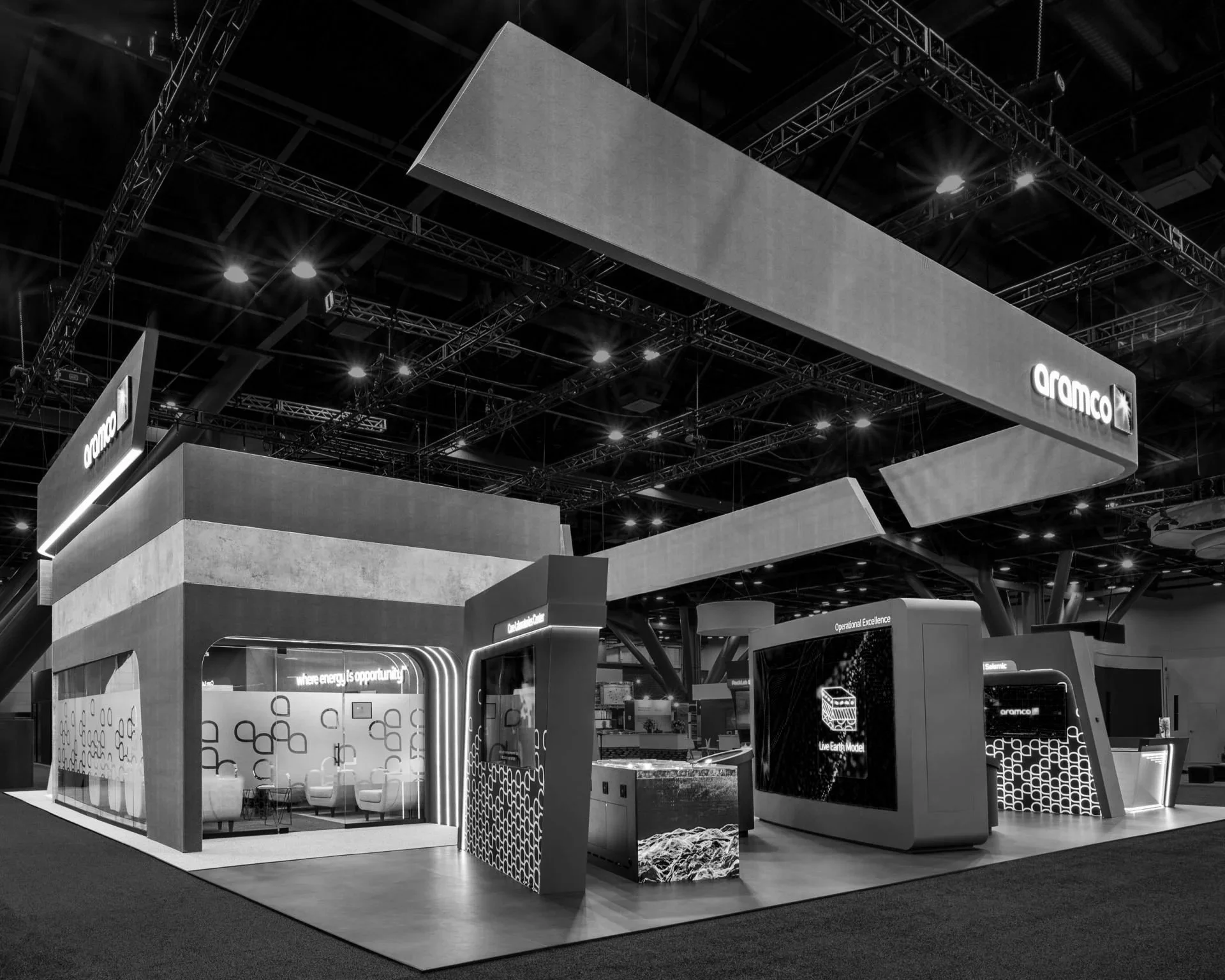 Black and white photo of an exhibition booth for a company called 'Aramco'. The booth features modern, geometric design elements, with a seating area, display screens, and a large overhead sign bearing the company's name. There are bright overhead lights and an open, spacious layout.