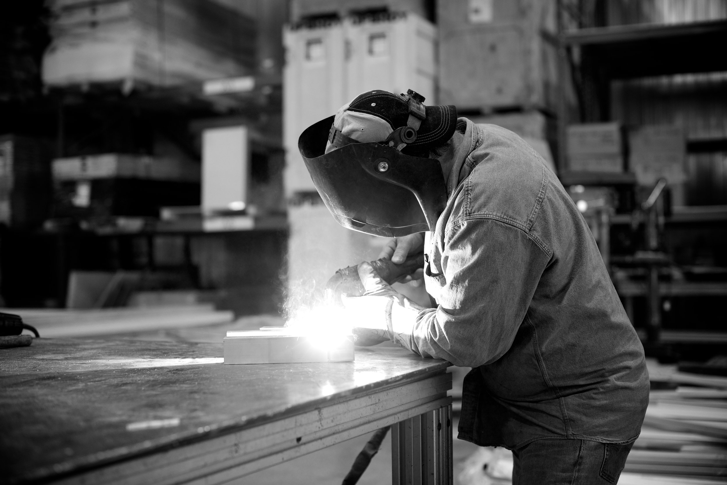 A person welding metal in a workshop, wearing protective gear including a welding helmet, gloves, and a long-sleeve shirt.