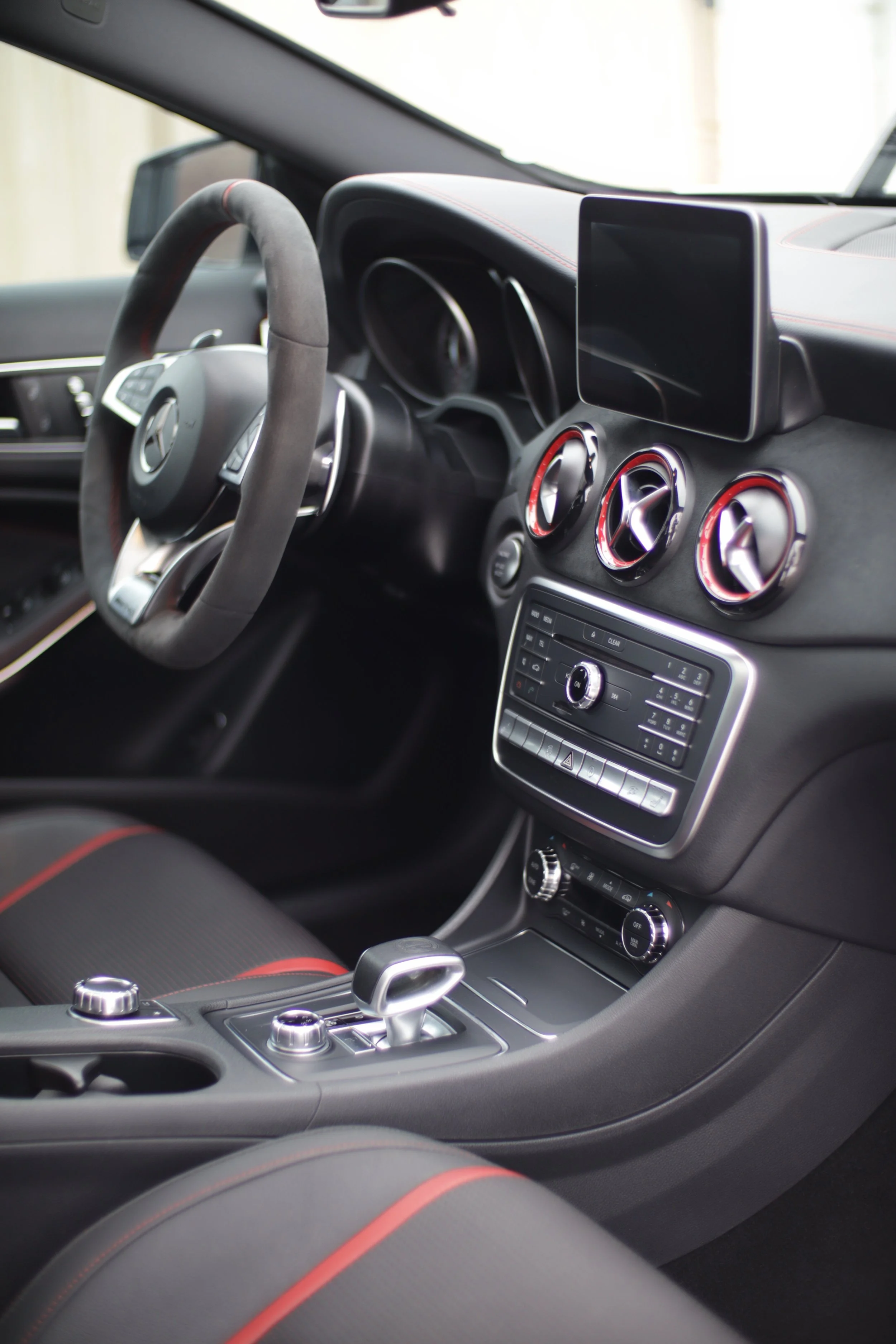 Interior of a modern Mercedes-Benz with Wild Detail car featuring a black dashboard, a multimedia screen, three circular air vents with red accents, control buttons, and a gear shifter.