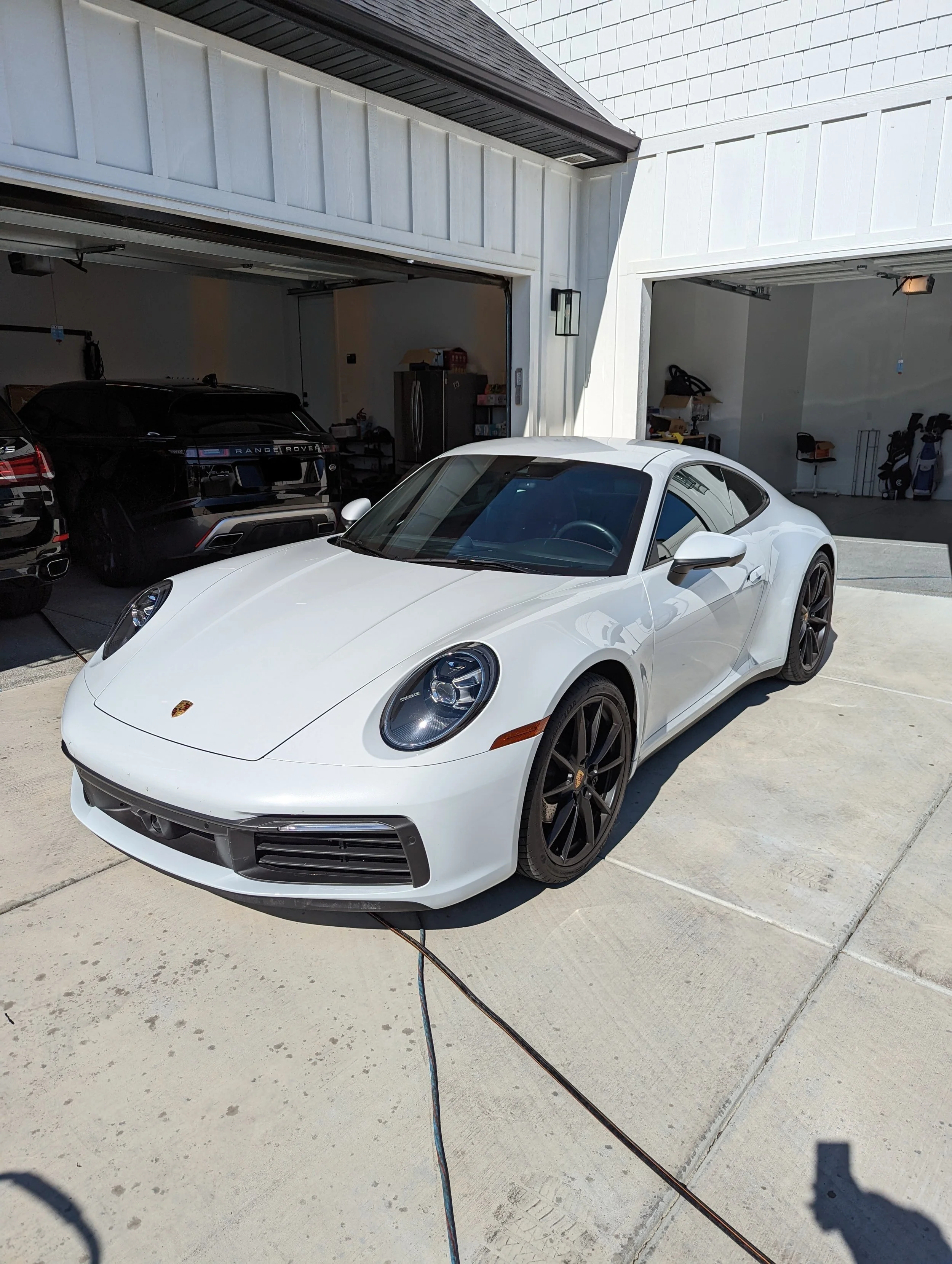 White Porsche sports car with Wild Detail parked in driveway with two garages in the background, one containing black cars and garage tools.