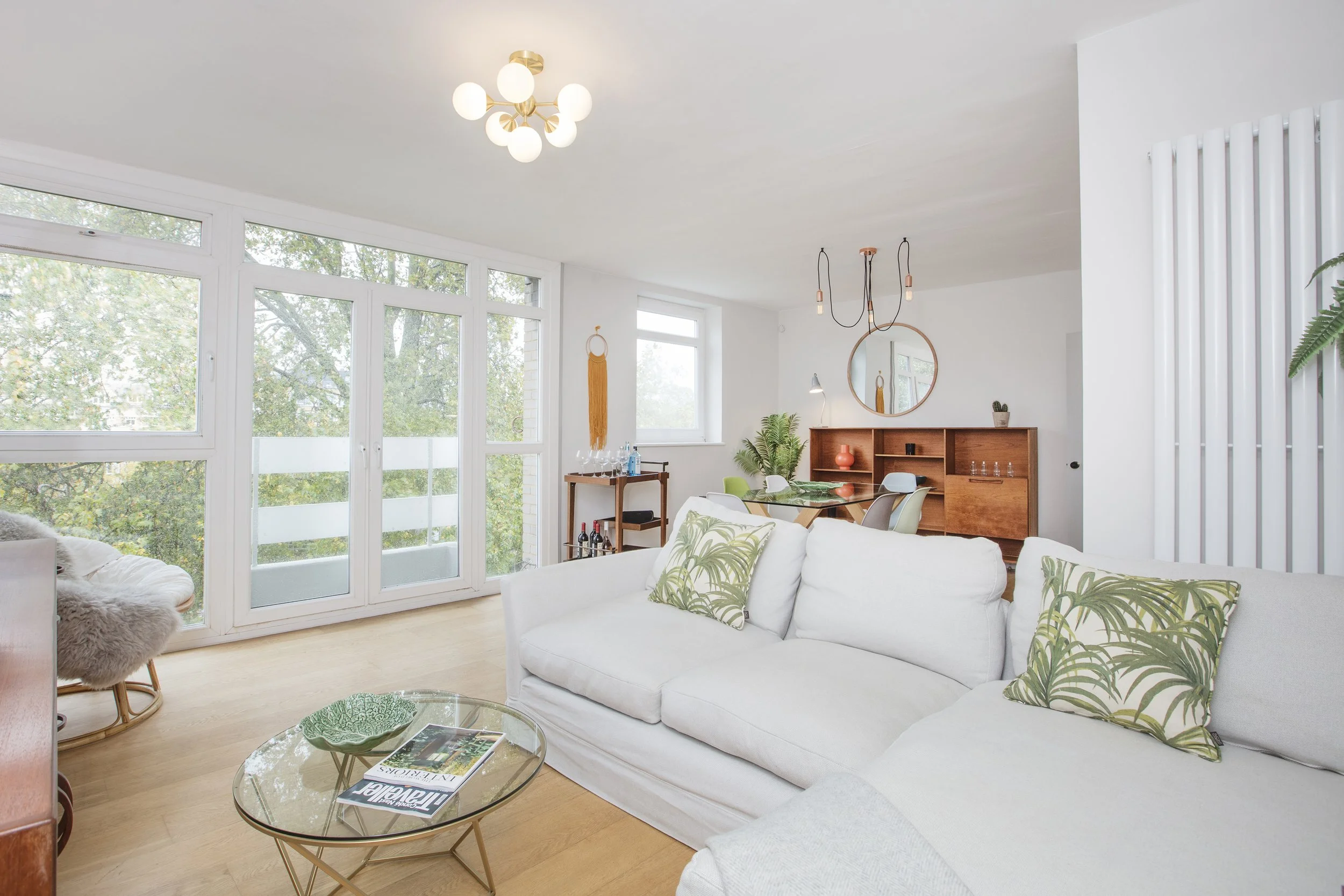 Modern living room interior with a white sofa, glass coffee table, and green accent pillows. Features a large window with tree view, wooden shelving, dining area, and stylish lighting. Decorative plants and accessories enhance the contemporary decor.