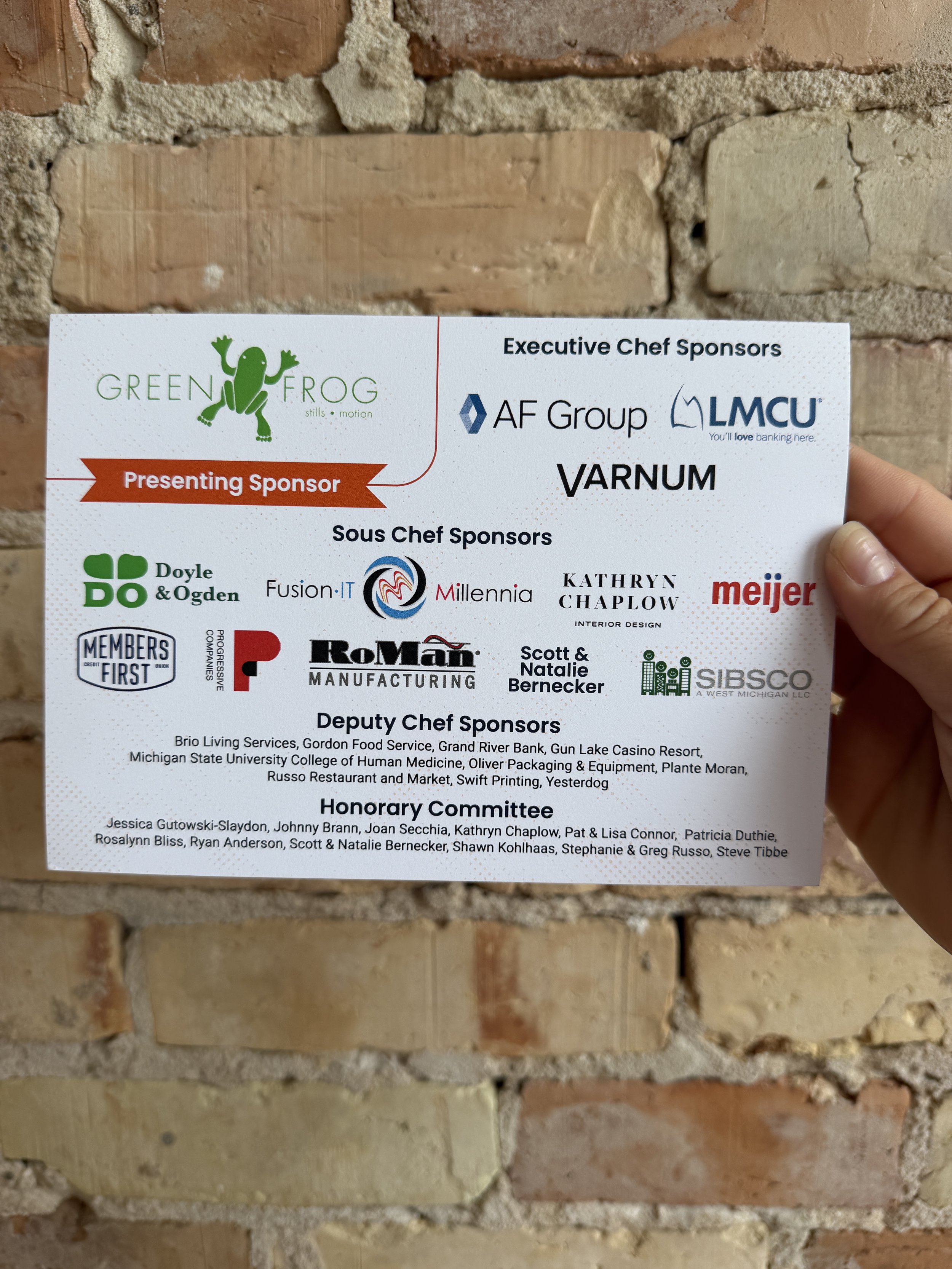 Brick background with a hand holding an invite with sponsor logos
