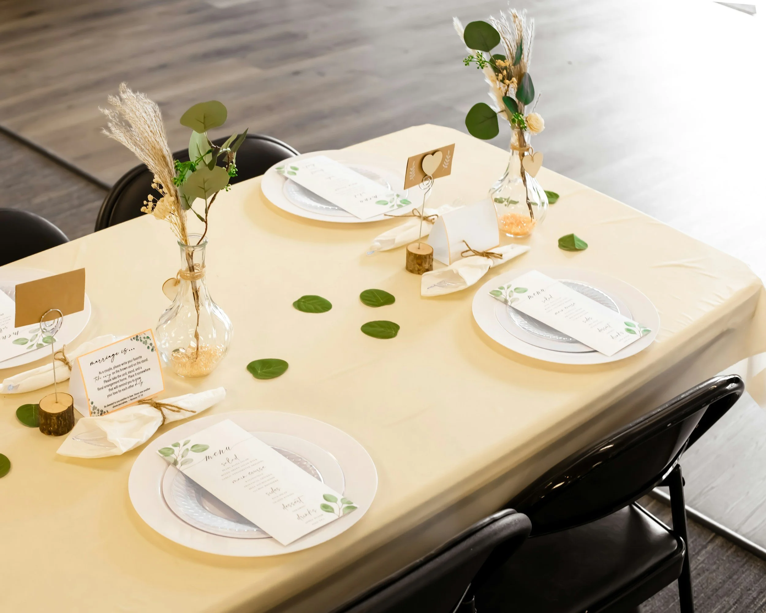Decorated wedding reception table with white tablecloth, greenery accents, glass vases with dried flowers, candles, and place settings including menus and napkins.