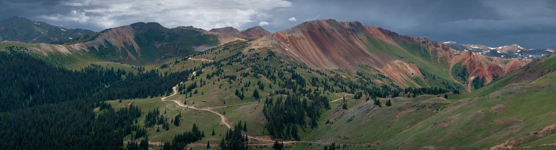 Mountain landscape with green forests and colorful, rugged mountain peaks under a cloudy sky.