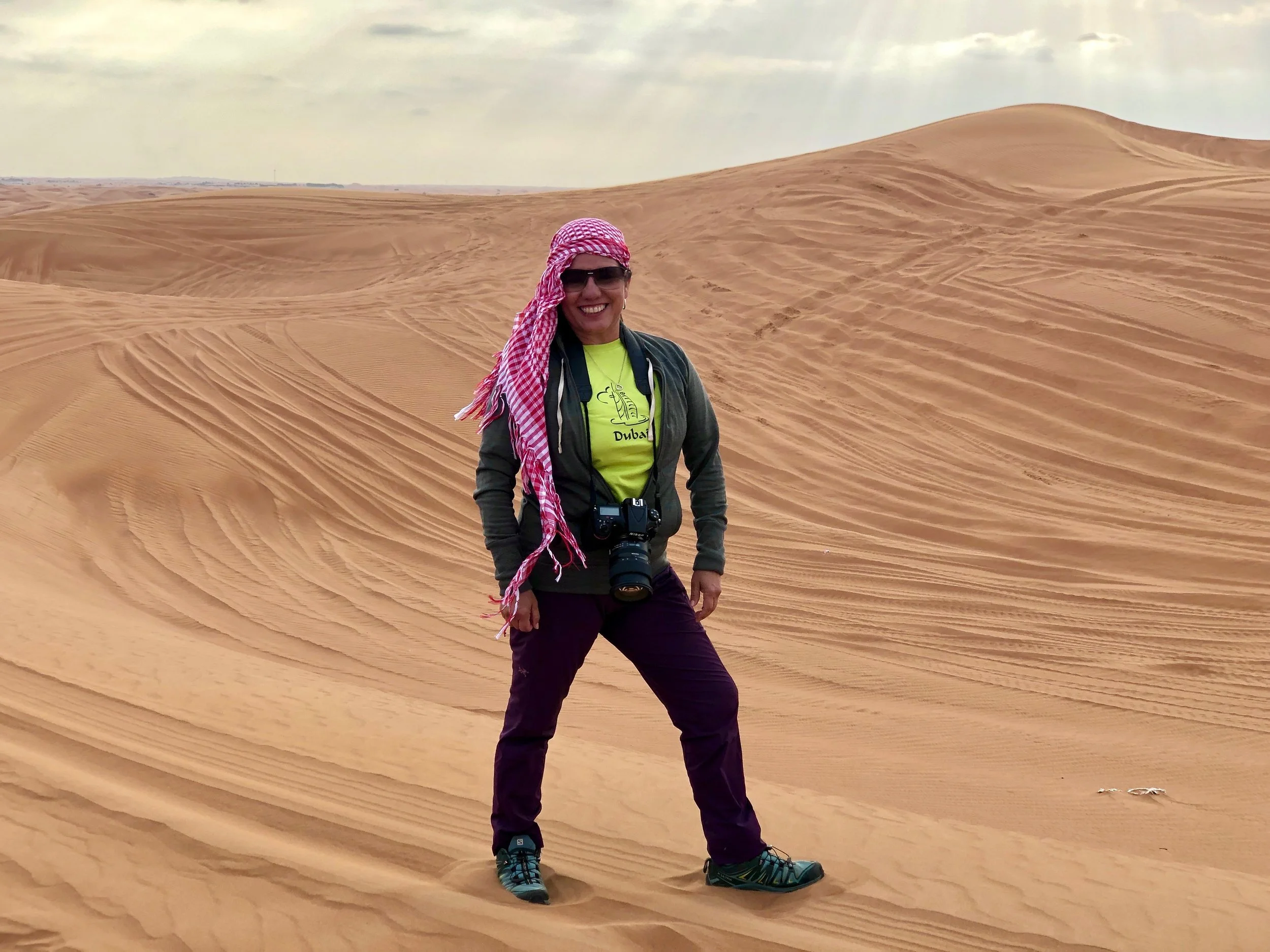 Person standing on sand dunes wearing a red and white headscarf, green jacket, and dark pants, with a camera around their neck.