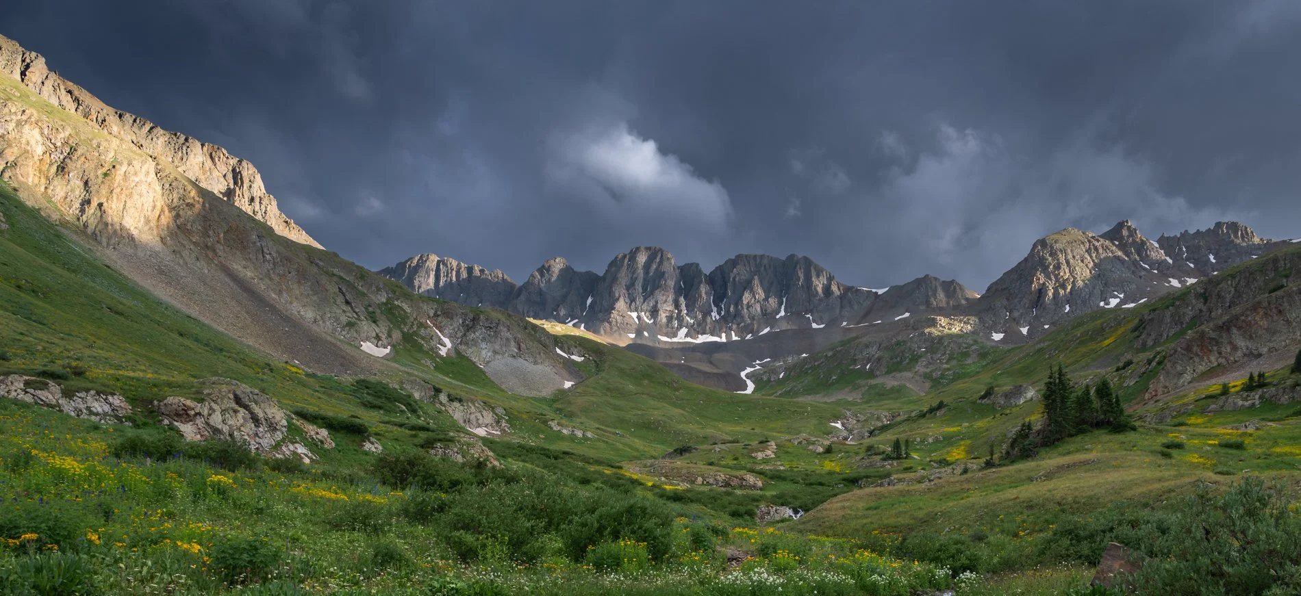 A scenic mountain landscape with green meadows, rocky slopes, and snow patches under a cloudy sky.