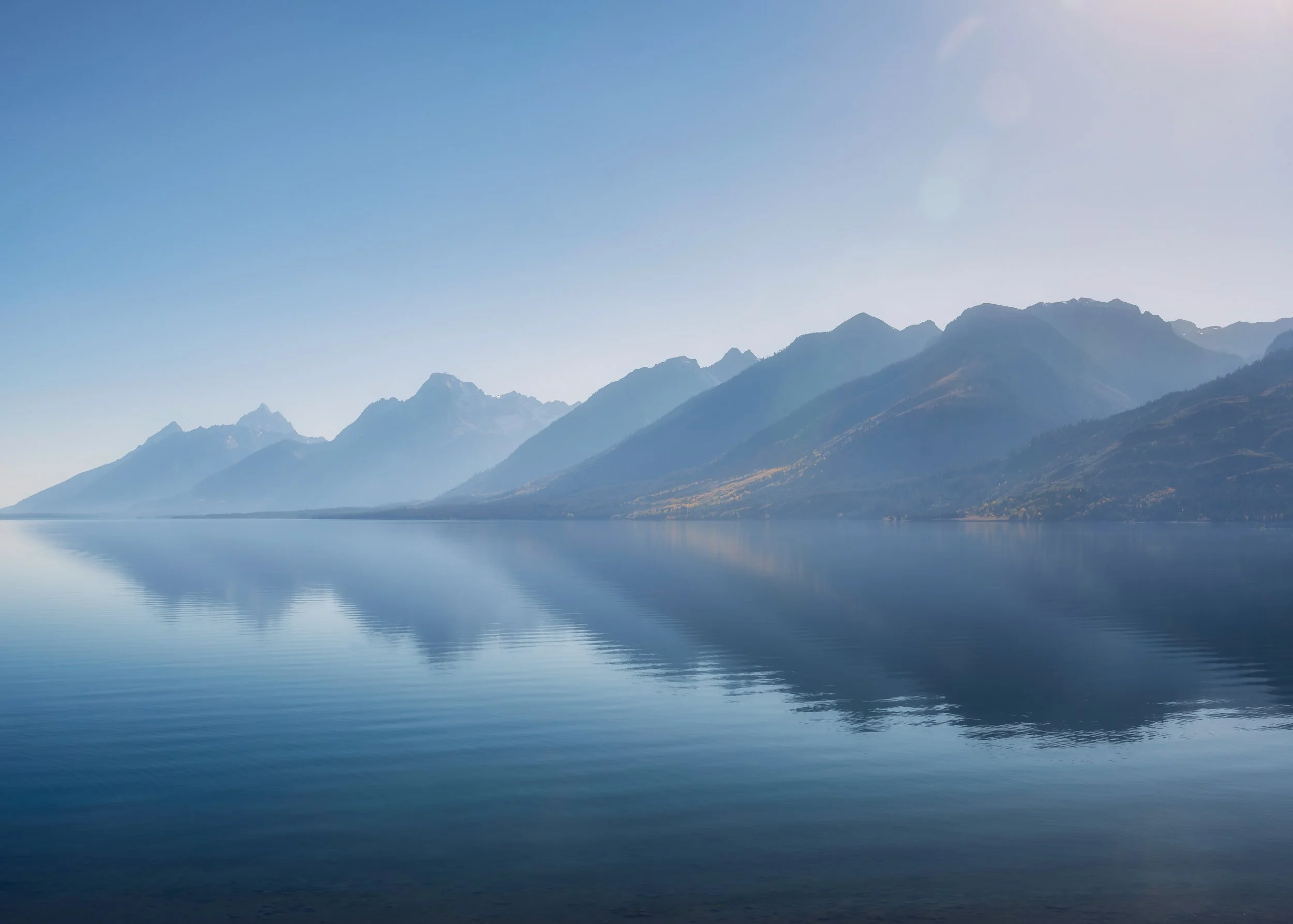 Wyoming Serene lake with calm water and a mirror-like reflection of distant mountain range under a clear blue sky.