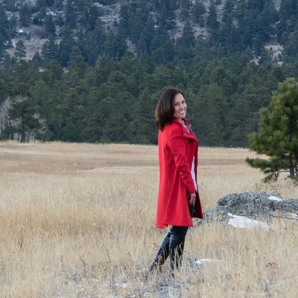 Woman in red coat standing in a grassy field with forested hills in the background.