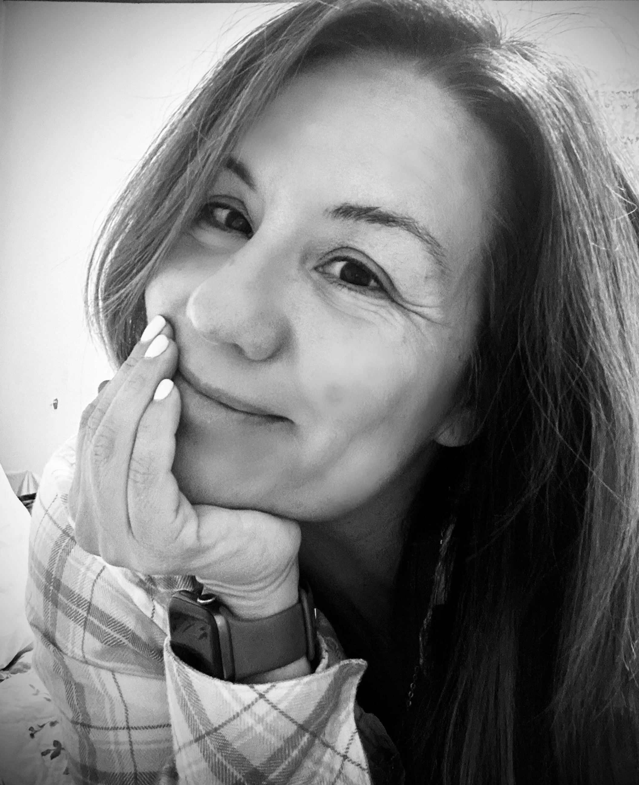 Smiling woman in a black and white portrait with long hair, wearing a plaid shirt and a smartwatch, resting her chin on her hand.