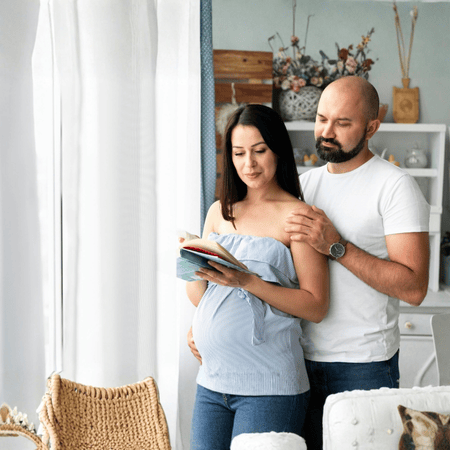 A pregnant woman reading a book with her partner standing behind her, holding her belly, in a cozy, well-decorated living room.