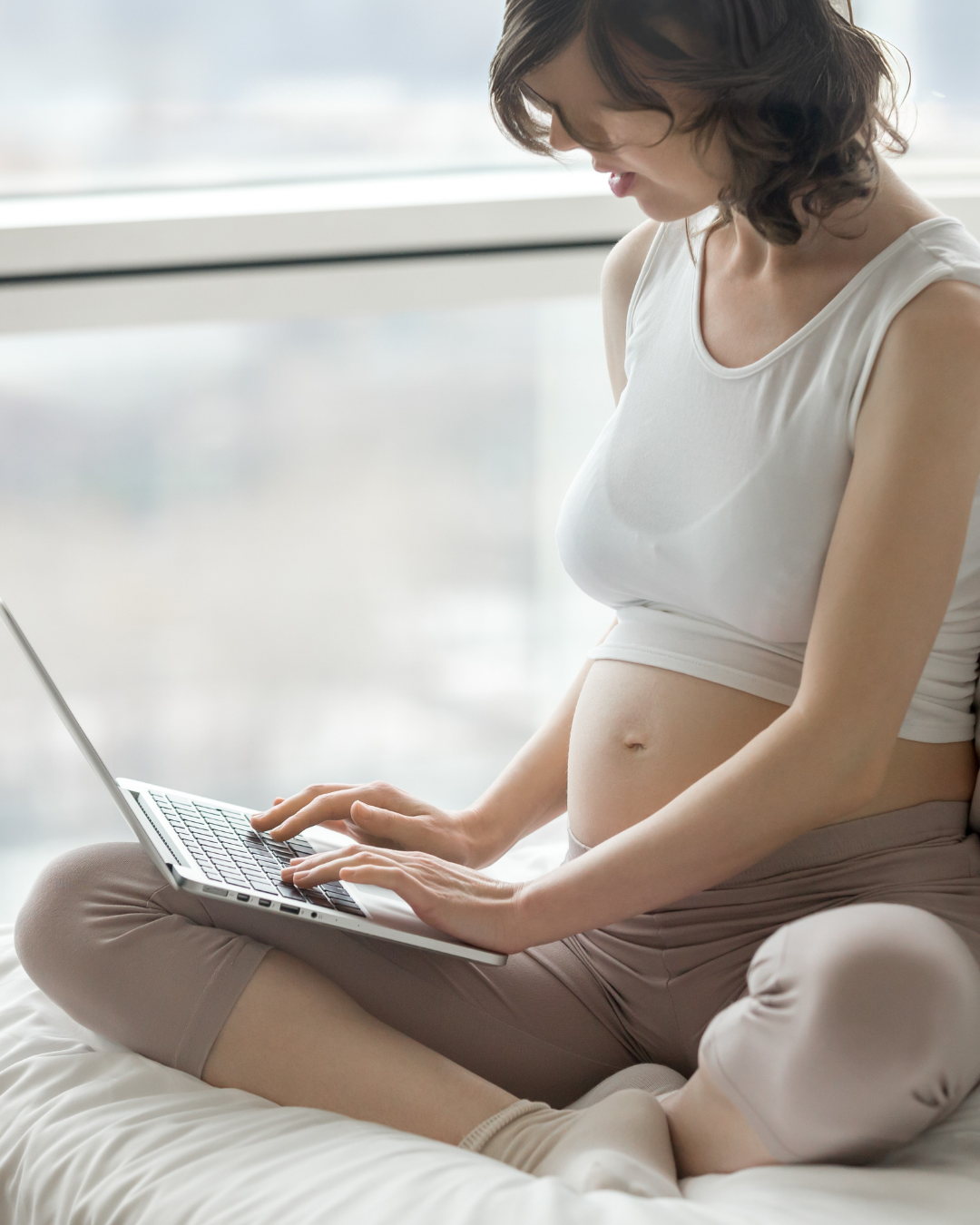 A pregnant woman sitting cross-legged on a bed and using a laptop in front of a large window with a view of a cloudy sky.