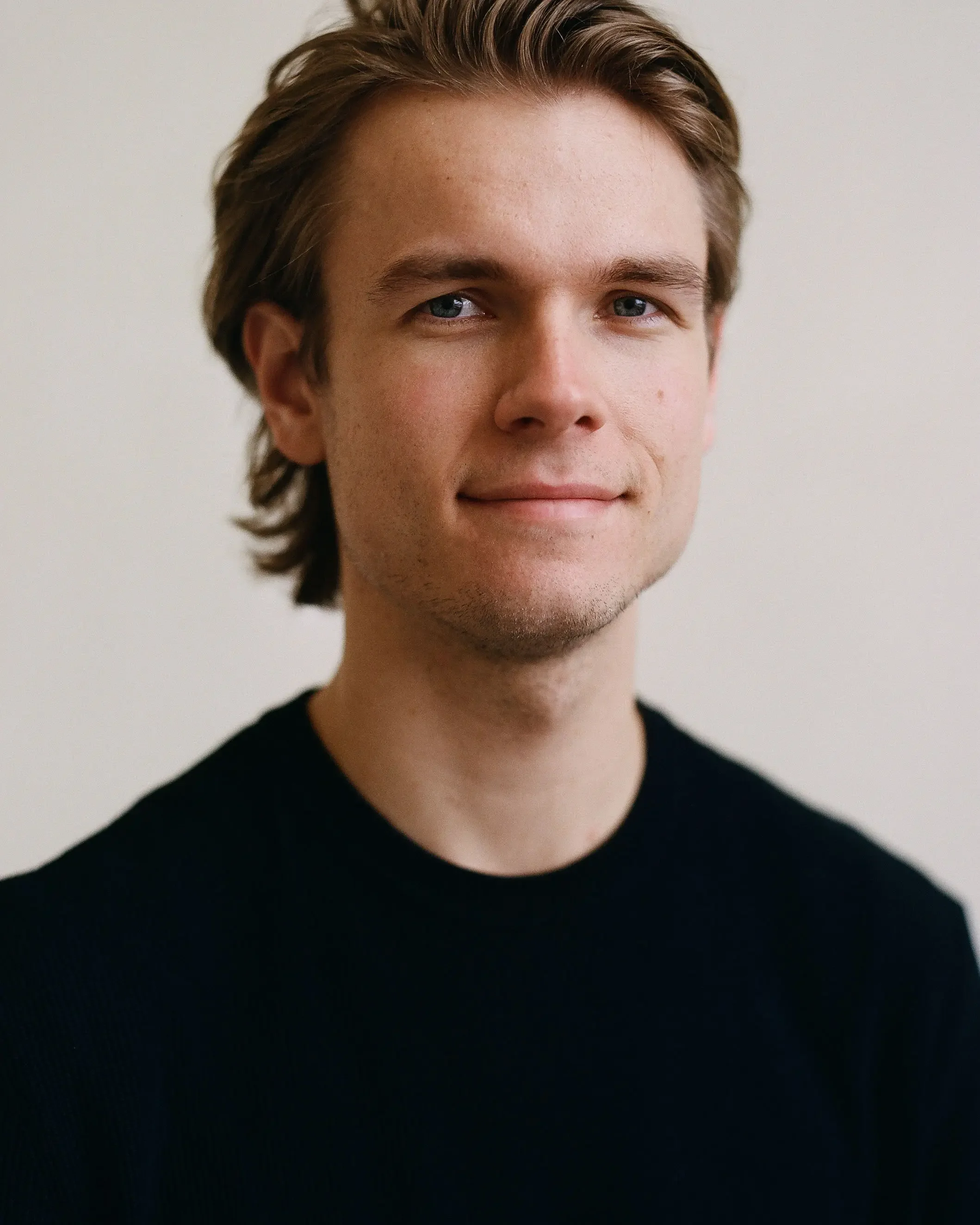 Close-up of a young man with light skin, blue eyes, and brown hair, wearing a black shirt, standing against a plain white background.