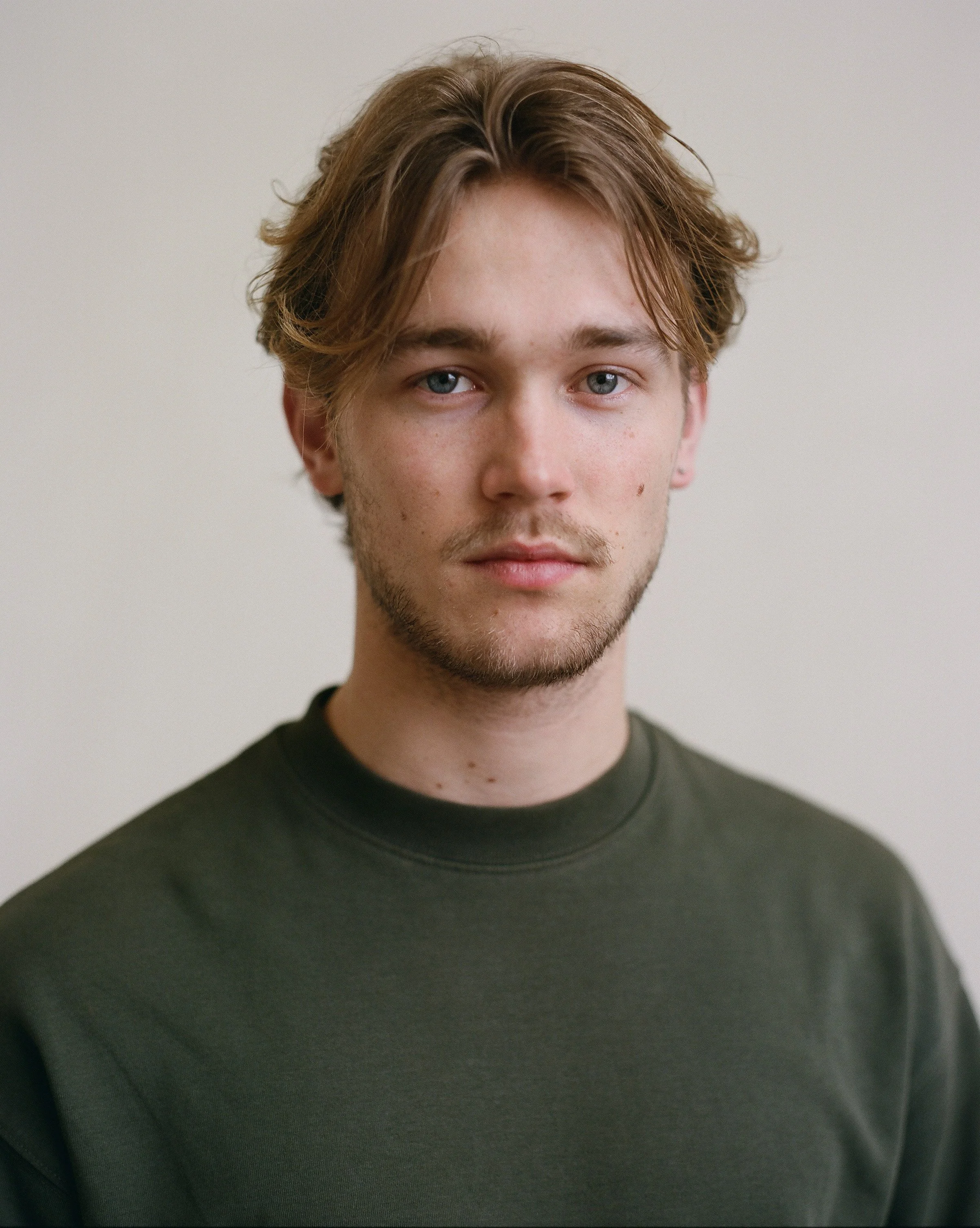 Close-up portrait of a young man with light skin, brown wavy hair, blue eyes, light beard, and freckles, wearing a dark crewneck shirt against a plain neutral background.