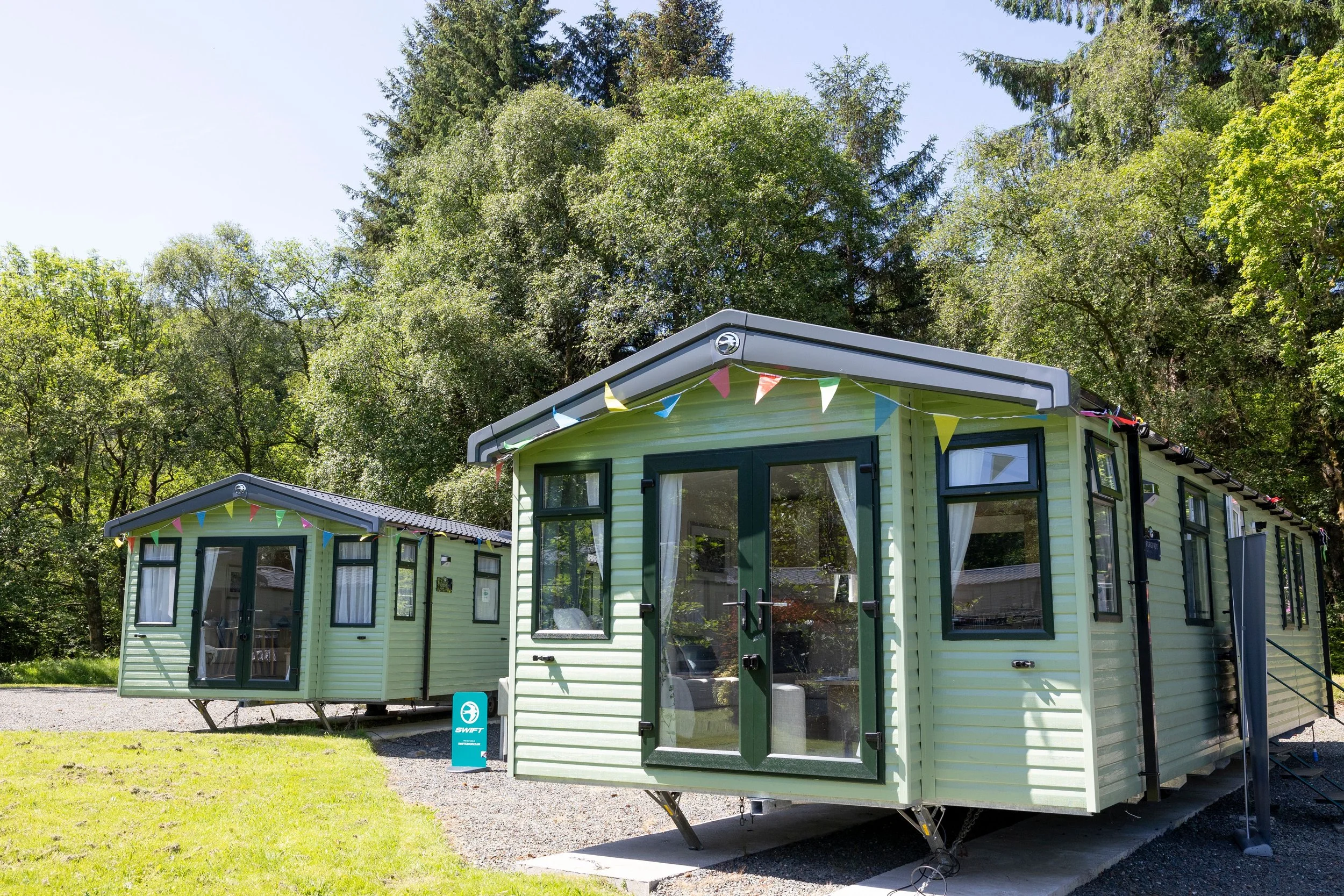 Sales Units at Inverbeg Holiday Park. Two green mobile homes with bunting and surrounded by tall trees on a sunny day.