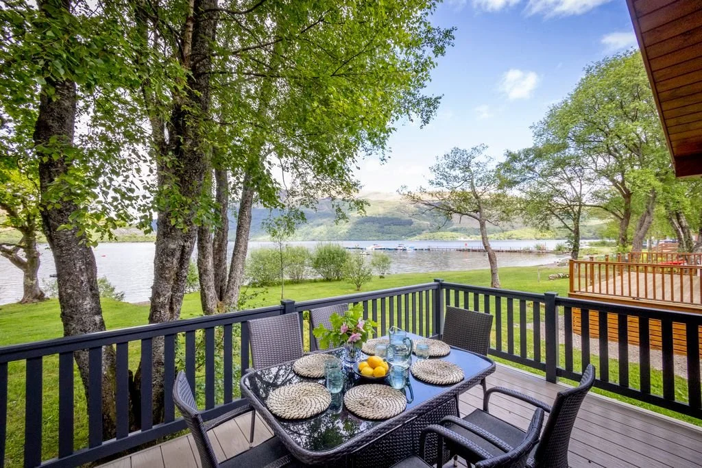 A lakeside outdoor dining area with a glass table set for six, surrounded by wicker chairs, overlooking a lush green yard, trees, and a lake with boats in the background.