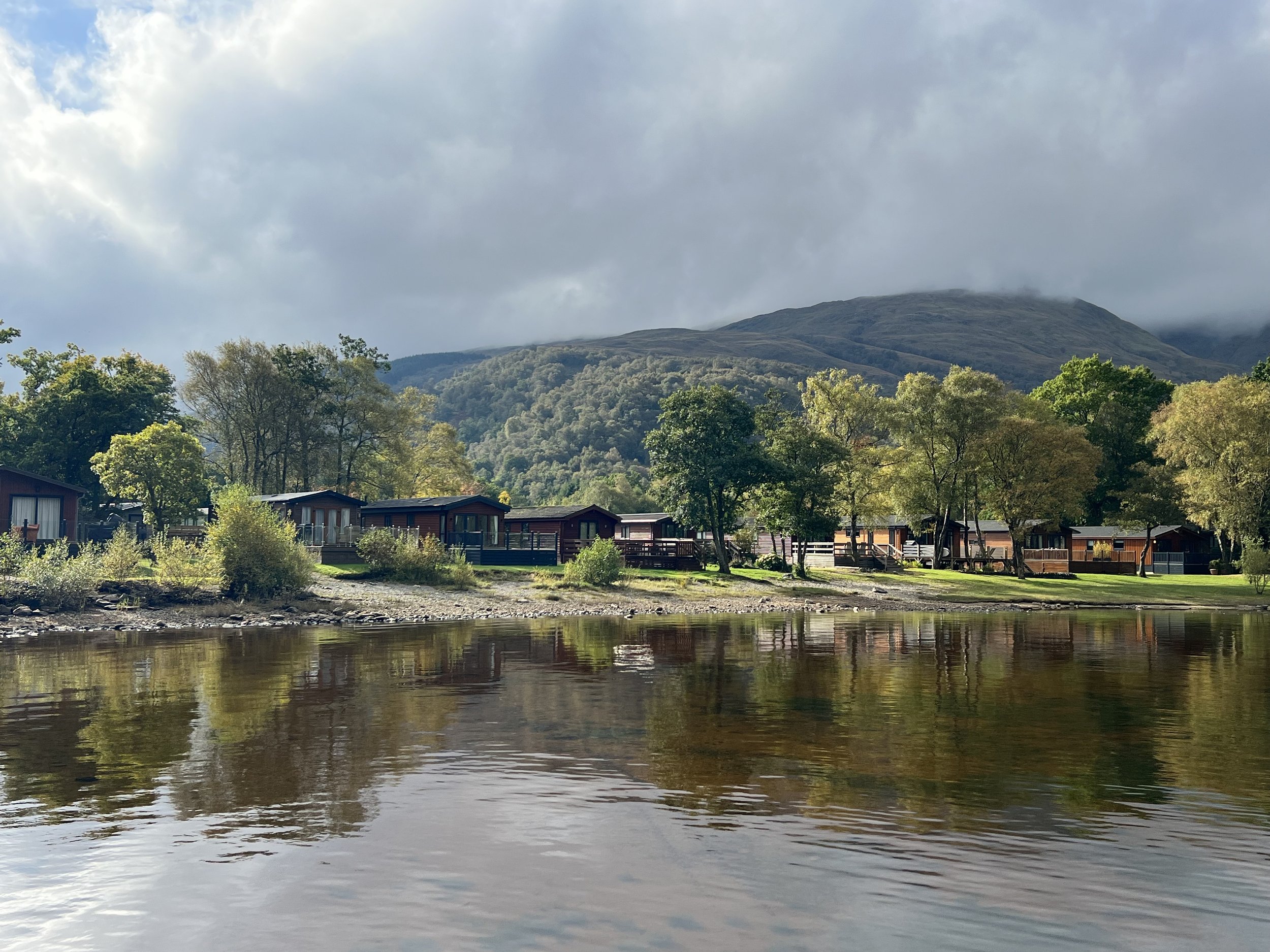 View from Pontoon at Inverbeg Holiday Park. Scenic view of Loch Lomond with reflections of trees and houses along the shore, surrounded by green hills and mountains under a cloudy sky.