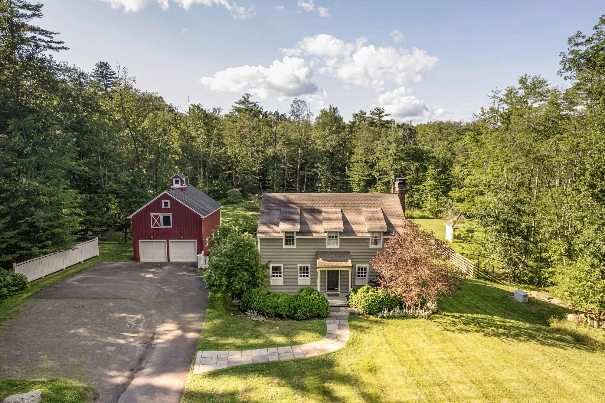 A large, two-story house with gray siding, surrounded by lush greenery and a well-maintained lawn. There is a curved concrete walkway leading to the front door, and a red barn with white trim and a garage door in the background to the left.