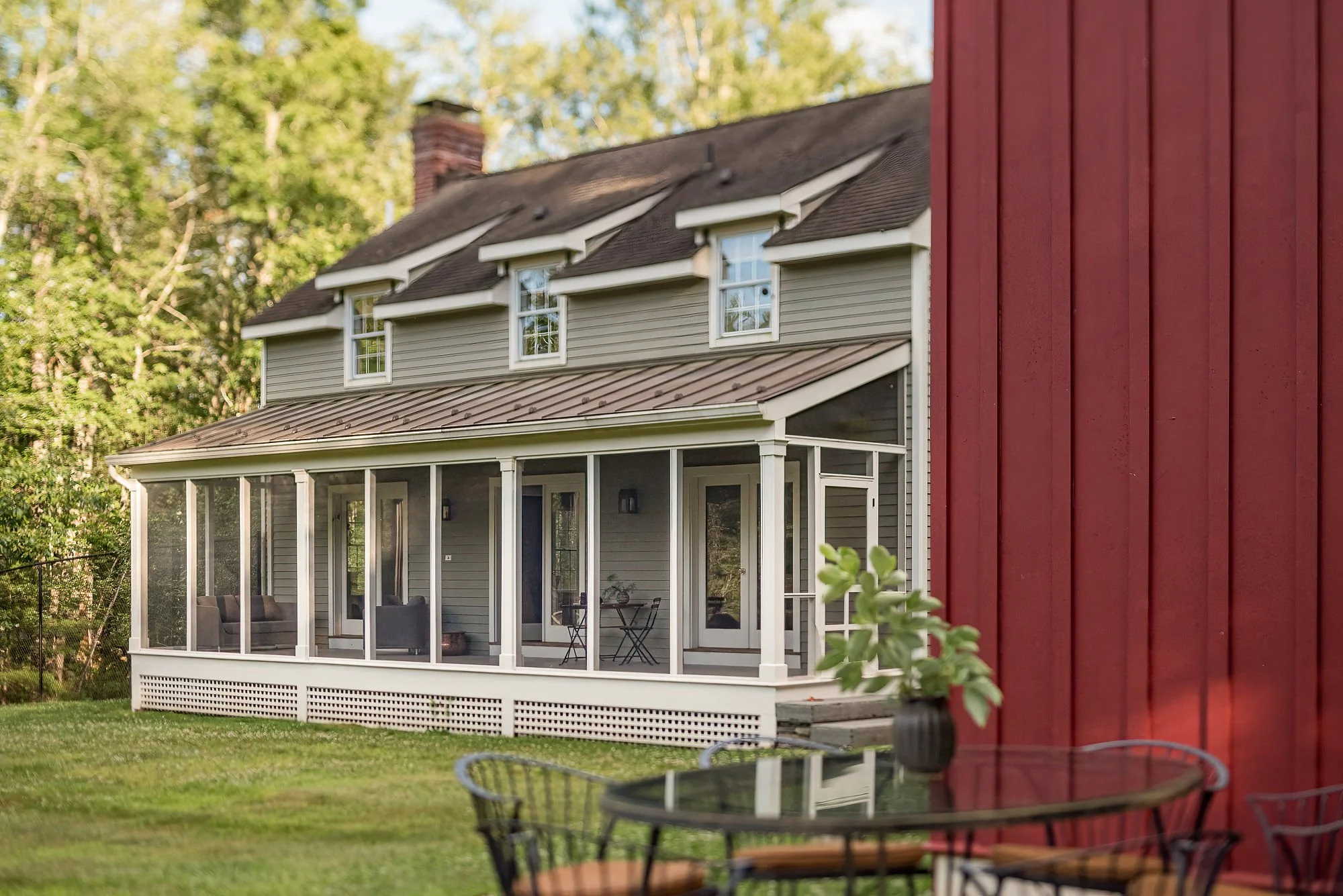 View of a house with a screened porch and a lawn in front, and a red building wall with a potted plant and outdoor table in the foreground.
