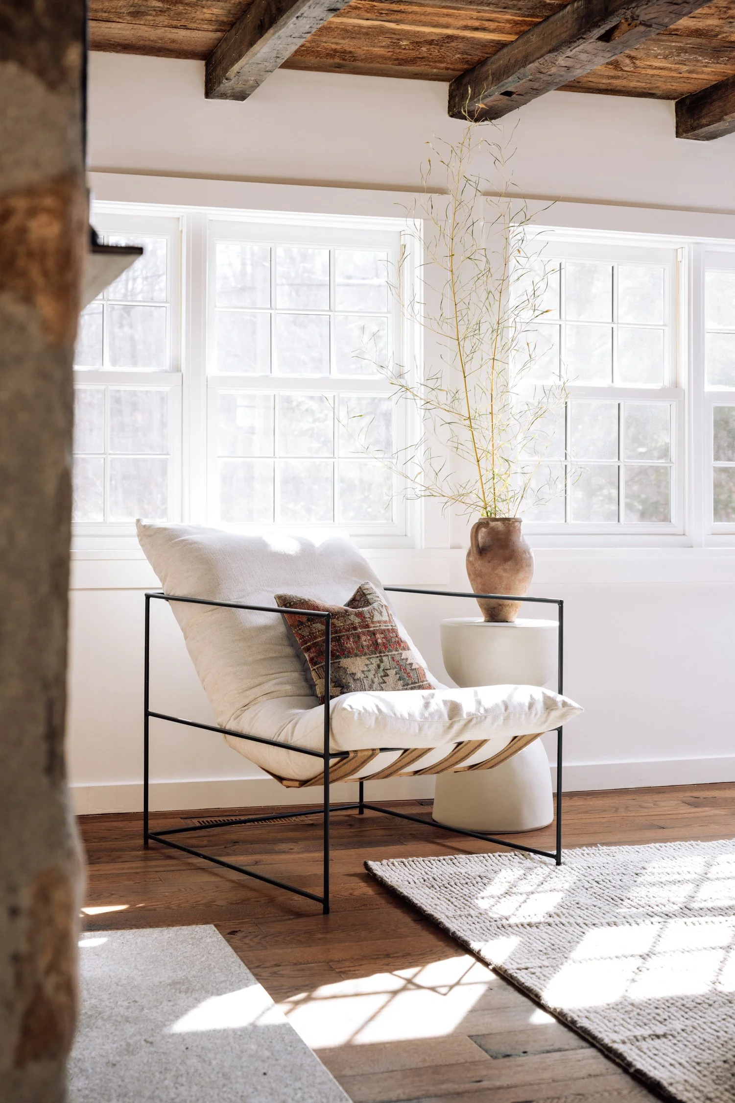 A cozy modern living room corner with a black metal frame lounge chair with white cushion and throw pillow, a white round side table with a large terracotta vase holding tall, thin green branches, large window with white trim, wooden ceiling beams, h