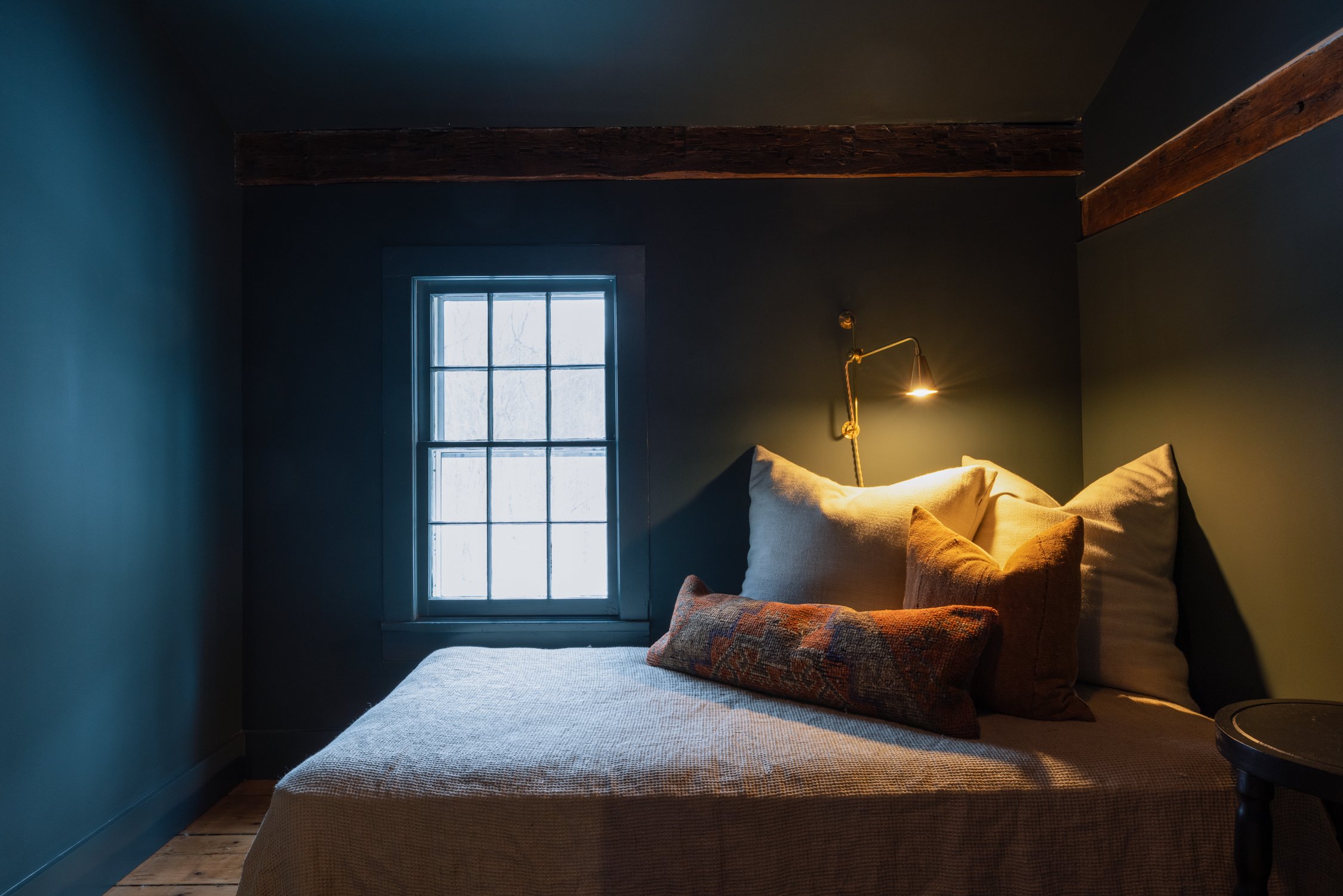 A cozy bedroom with dark green walls, a window with grid panes, a bed with beige and patterned pillows, and a wall-mounted reading lamp.