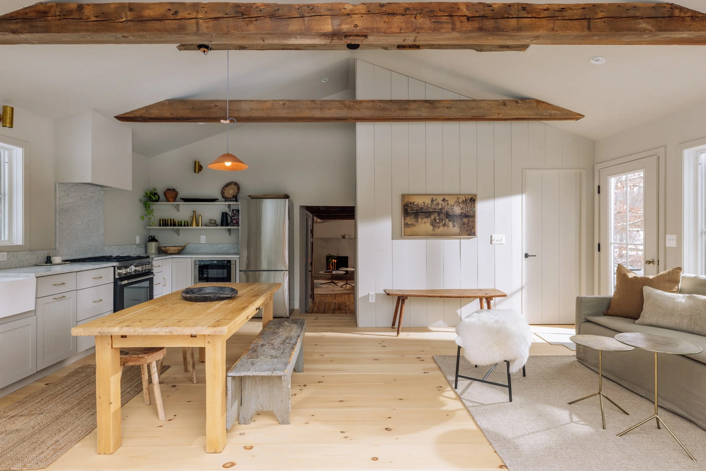 Open-concept kitchen and living room with wooden flooring, exposed wooden beams, white walls, and natural light from windows and a glass door, featuring a wooden dining table, a gray sofa, and a decorated shelf.