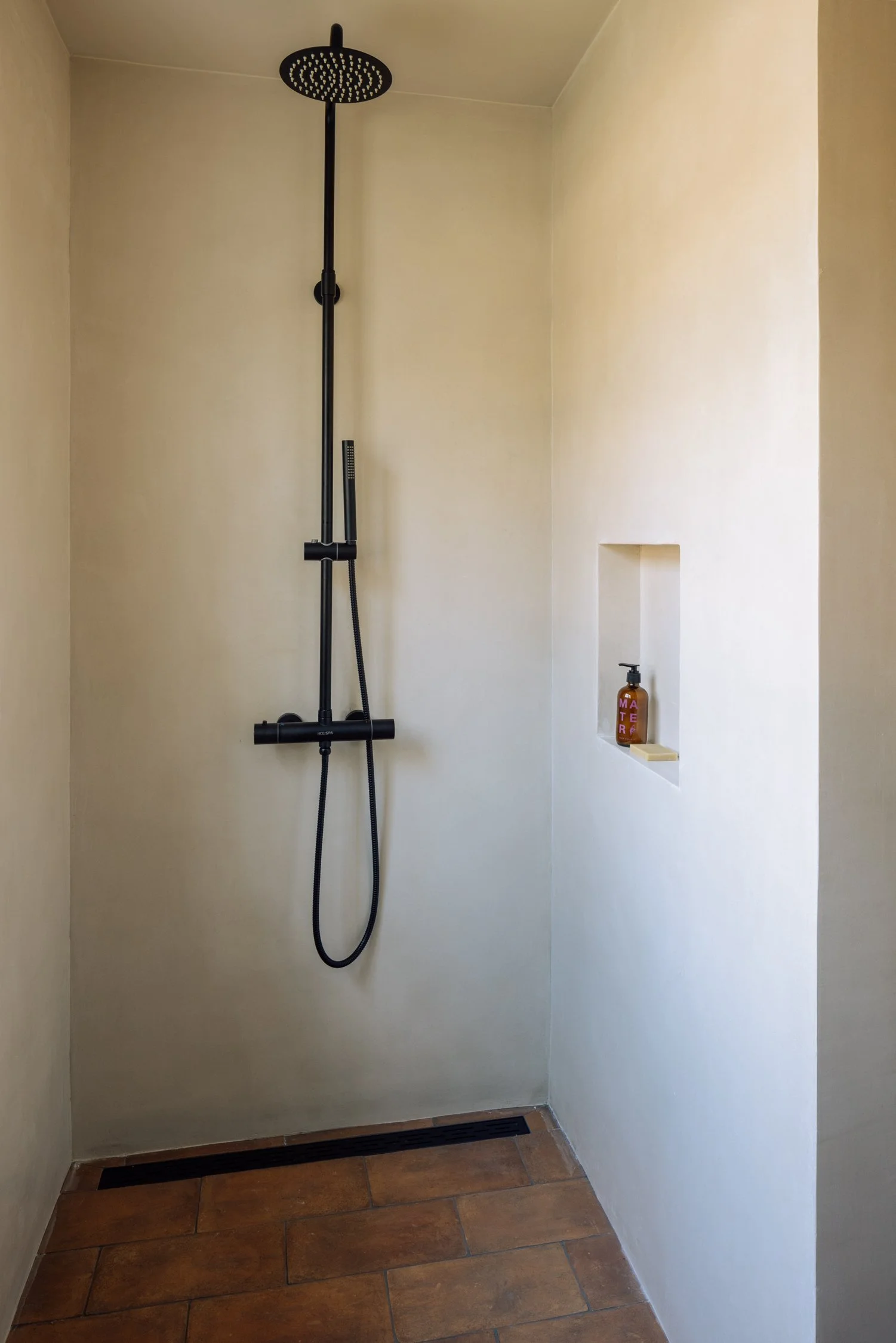 Shower with a black rain showerhead, handheld shower, and wall niche containing soap and hand sanitizer in a neutral bathroom.