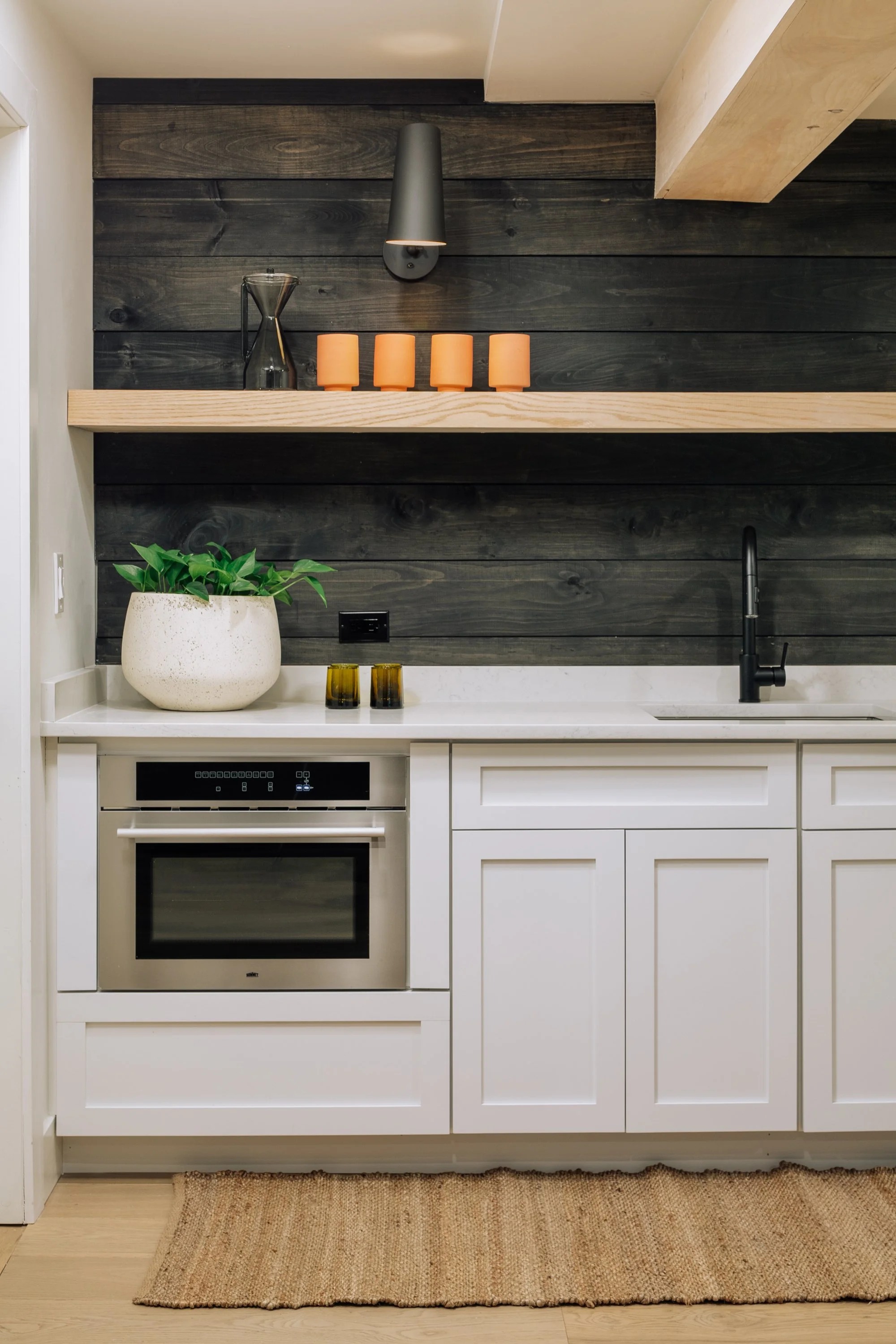 Modern kitchen with white cabinetry, black faucet, black wall-mounted light, dark wood panel wall with a floating wooden shelf holding orange cups, a glass pitcher, and a black vase, and a potted plant on the counter.