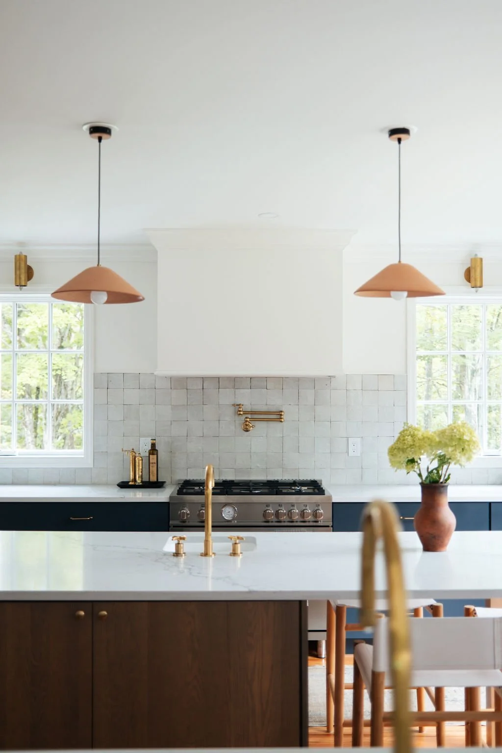 Modern kitchen with white walls, large windows, black lower cabinets, and a white marble island with gold faucet. There are two hanging pendant lights with tan shades, a stove with brass fixtures, and a flower vase with yellow flowers on the counter.